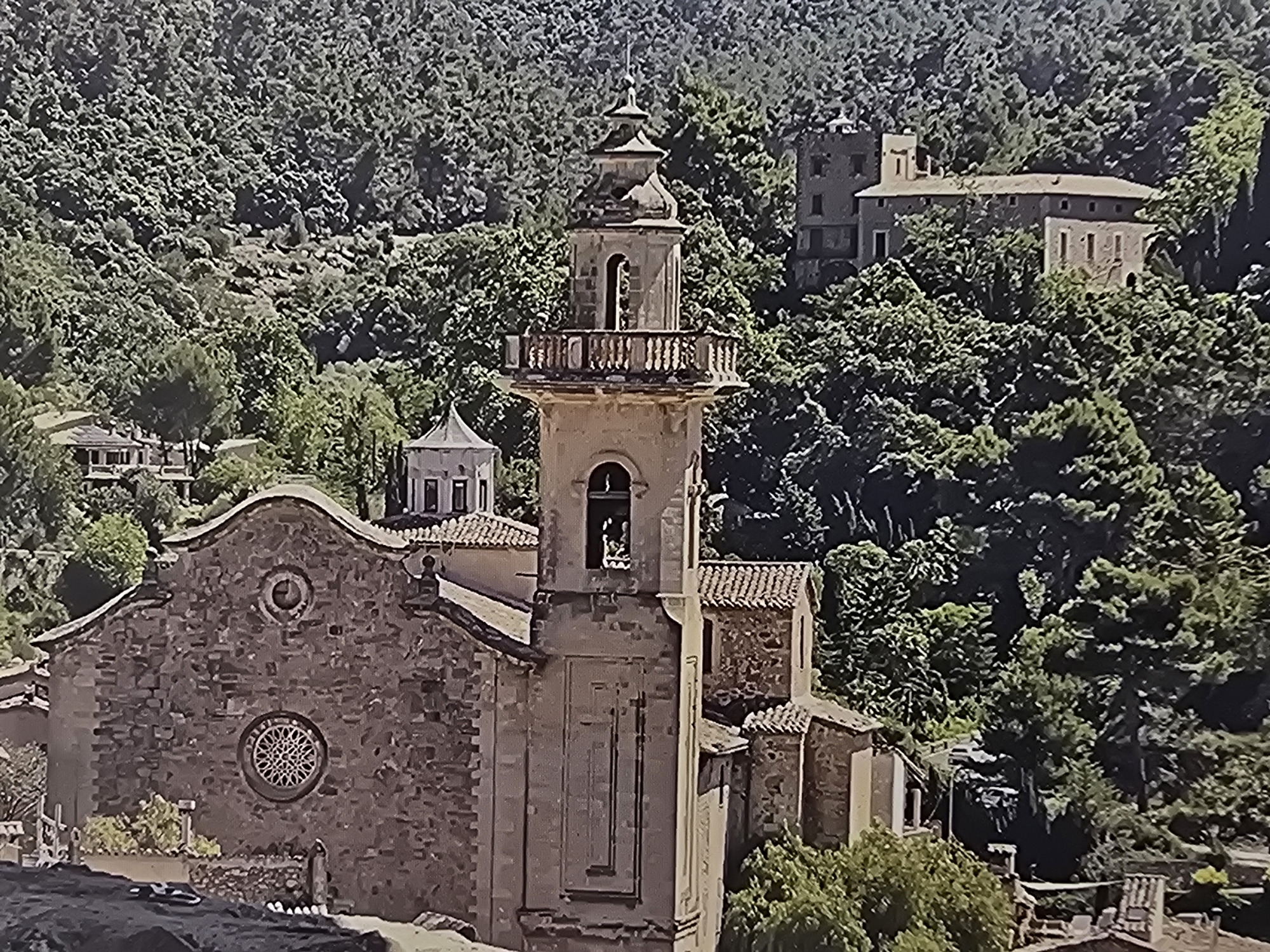 The central subject of the image is a stone church featuring a prominent bell tower with a balustraded upper level and a small domed spire. The church facade exhibits a large, ornate rose window and a smaller circular window above it. Behind the church, a building with a light-colored dome is partially visible. Further up the densely forested hillside, an older, substantial stone building or complex, possibly a manor or monastery, with multiple windows and a tower, is nestled among the trees. The scene is dominated by lush, green, forested mountainsides that rise steeply behind the structures. No people are visible.
No specific activity or event is taking place; the scene is static.
The image depicts a serene, historical village or settlement nestled within steep, verdant hills. The architecture, characterized by robust stone construction, terracotta roof tiles, and the style of the bell tower and windows, suggests a European, likely Mediterranean, region. The time of day appears to be bright daytime, with clear visibility and ample sunlight illuminating the buildings and foliage. The weather is clear and sunny. The dense forestry indicates a temperate or subtropical climate with abundant vegetation.
No visible text is present in the image.