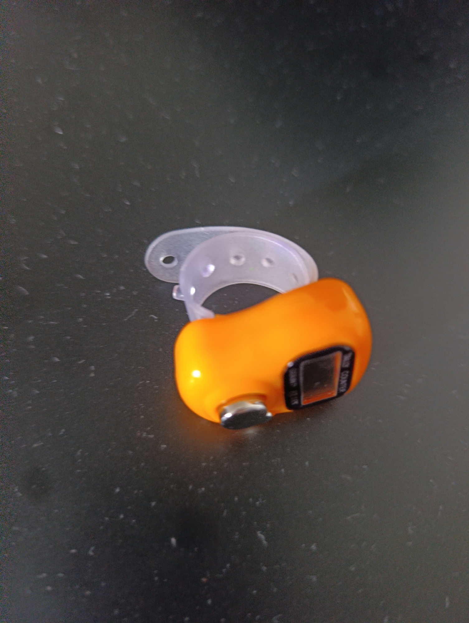 An orange digital prayer counter with a clear plastic ring is positioned on a dark, speckled surface. The prayer counter is small and has a digital display. The clear plastic ring is a loop with several small holes along its surface. The surface the objects are on appears to be a tabletop. The lighting suggests an indoor environment, possibly a desk or counter. The image is taken from a slightly elevated angle.