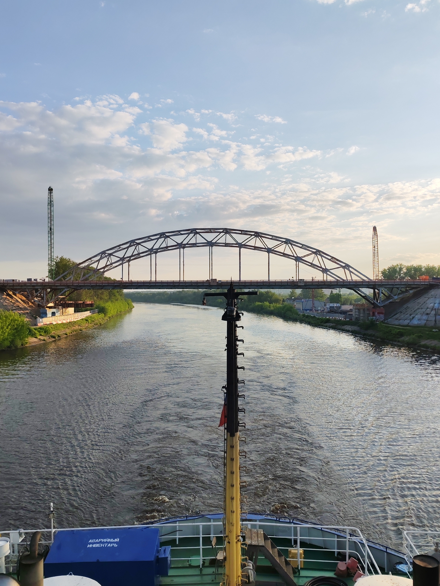 The image depicts a view from a ship sailing on the Moscow Canal in Dmitrov, Russia, under the Dmitrov Highway Bridge. The bridge is a steel arch structure with a road deck suspended below. Construction cranes are visible on either side of the bridge, indicating ongoing work. The canal is flanked by green vegetation and some buildings. The sky is partially cloudy with blue patches. The foreground shows the deck of the ship with a mast and other equipment. There is a blue box labeled in Russian "АВАРИЙНЫЙ ИНВЕНТАРЬ", which translates to "Emergency Equipment".