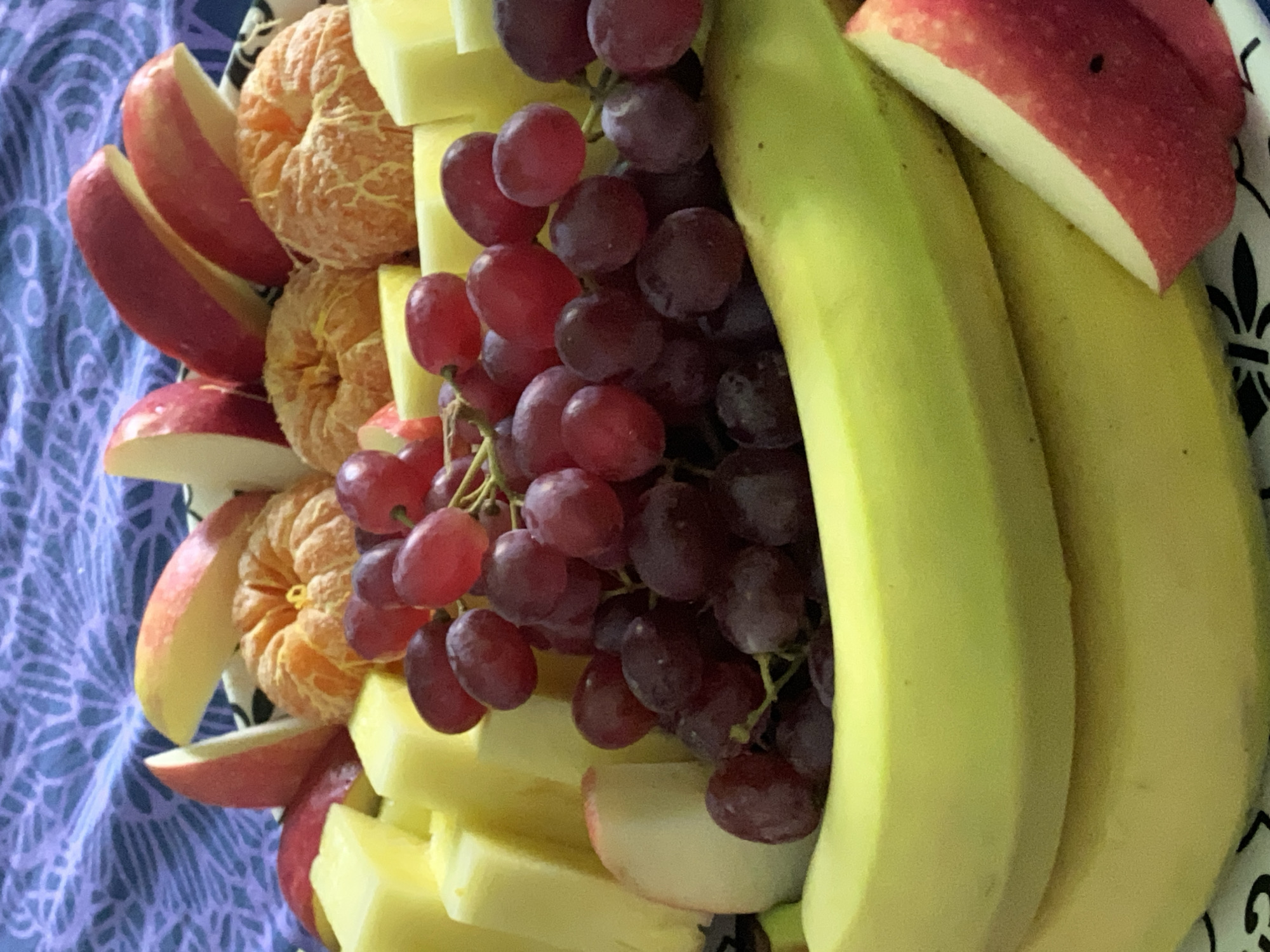 A top-down shot reveals a fruit platter composed of sliced red apples, peeled tangerines, cubed light yellow cheese, dark red grapes, and two ripe bananas. The platter rests on a blue and white patterned surface. The image doesn't provide sufficient information to establish a location in the city of Chaikovsky, Russia.