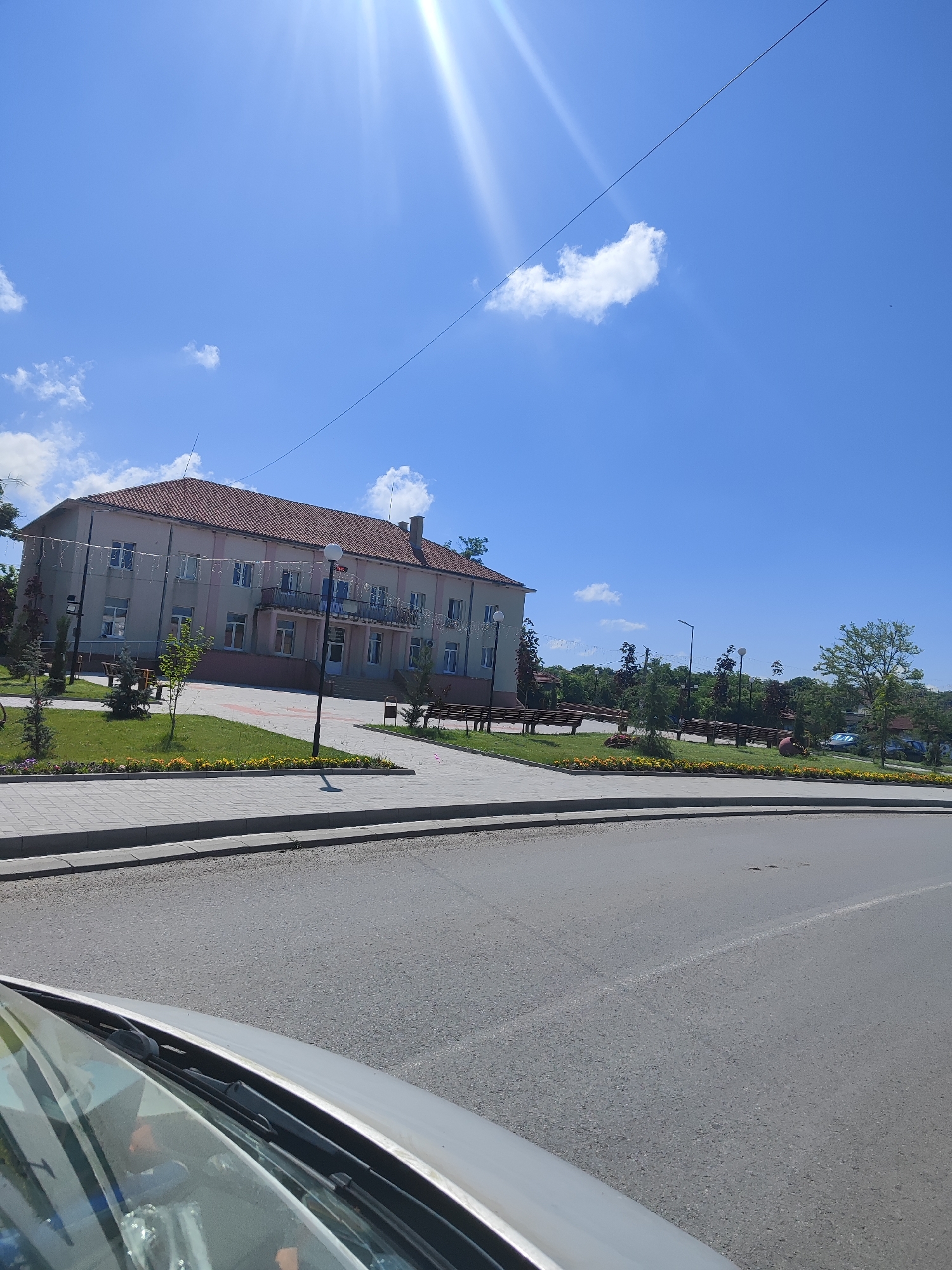 A low-angle, eye-level medium shot captures a sunlit exterior scene in General Inzovo, Bulgaria. The sky is a brilliant blue with scattered white clouds and a lens flare from the sun. In the middle ground, a long, two-story building with a red tile roof stands prominently. The building has numerous windows and a central balcony supported by columns. Decorative lights are strung along the facade of the building. In front of the building, a paved walkway borders a grassy area with small trees and park benches. A lamppost stands near the walkway. The foreground shows a section of asphalt road and the hood and windshield of a silver vehicle. The scene depicts a public space bathed in bright sunlight, suggesting a pleasant, clear day.