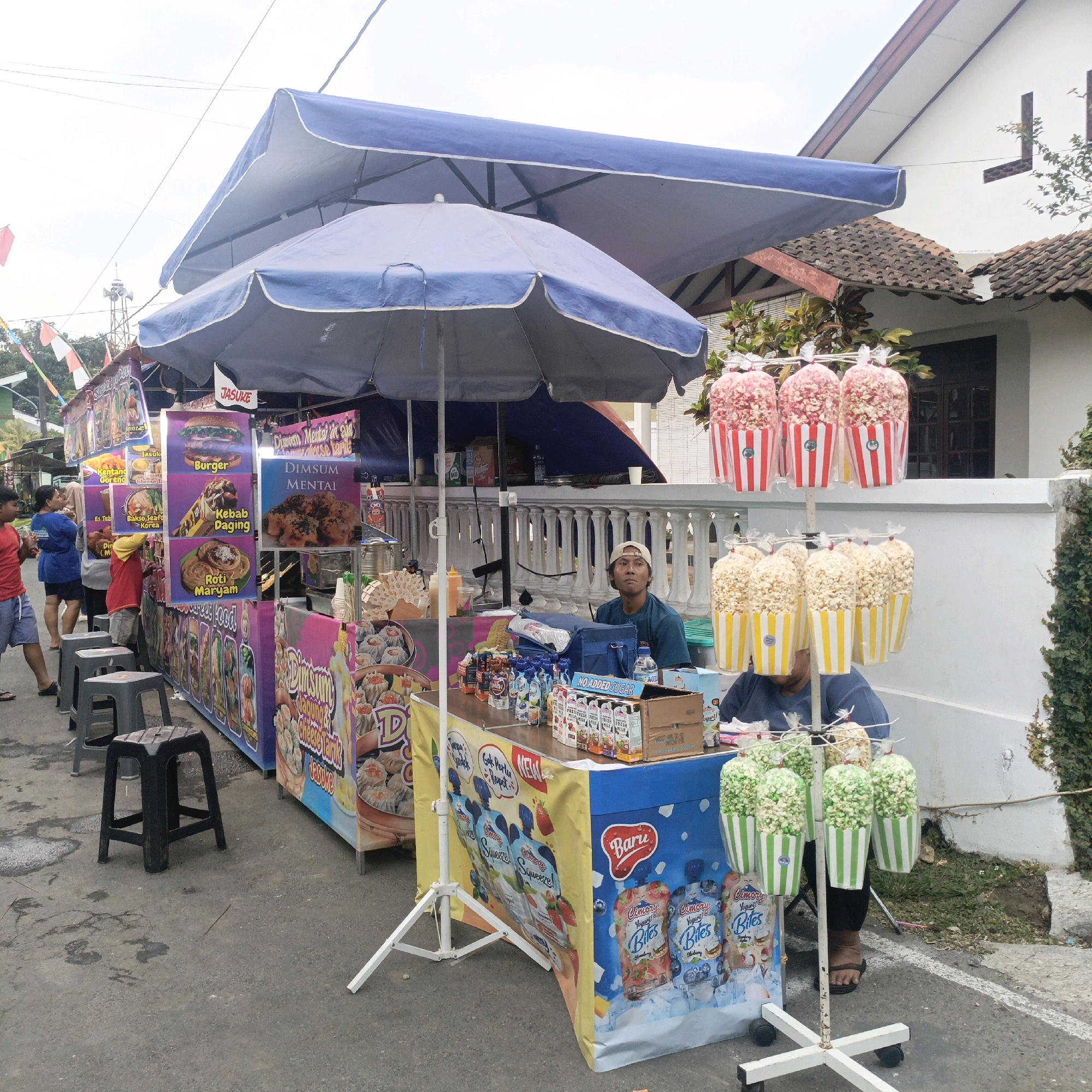 The image depicts a street food stall in Salatiga, Indonesia, with an outdoor setting. Two food vendors, both men, are visible. One vendor, wearing a blue t-shirt and a light-colored cap, is seated behind a table laden with drinks and snacks. The second vendor, wearing a red t-shirt and a yellow headscarf, is partially visible assisting customers.
The stall is organized with multiple sections selling various food items. Prominent signage displays images of burgers, kebabs, dim sum, and rotis. Bags of popcorn, some with red and white stripes and others with yellow and white stripes, are displayed on a rack, with both red and green colored popcorn visible. Various beverages, including bottled water and juice pouches, are also available. Several black stools are placed in front of the stall, suggesting a seating area for customers. The background shows a residential building with a tiled roof, a white wall, and some greenery. The ground is paved with asphalt, and a white line indicates a road boundary. Decorative flags are also visible in the distance. The overall scene suggests a lively street market or food fair.