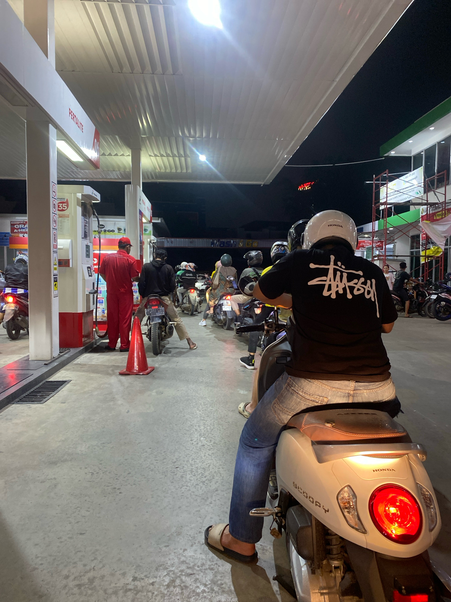 The image depicts a nighttime scene at a Pertamina gas station in Batam, Indonesia, with several people on motorcycles lined up to refuel. A gas station attendant in a red uniform is visible near a fuel pump. The ground is concrete, and there is an overhead canopy illuminated by lights. The overall atmosphere suggests a busy, ordinary evening at a common urban amenity.