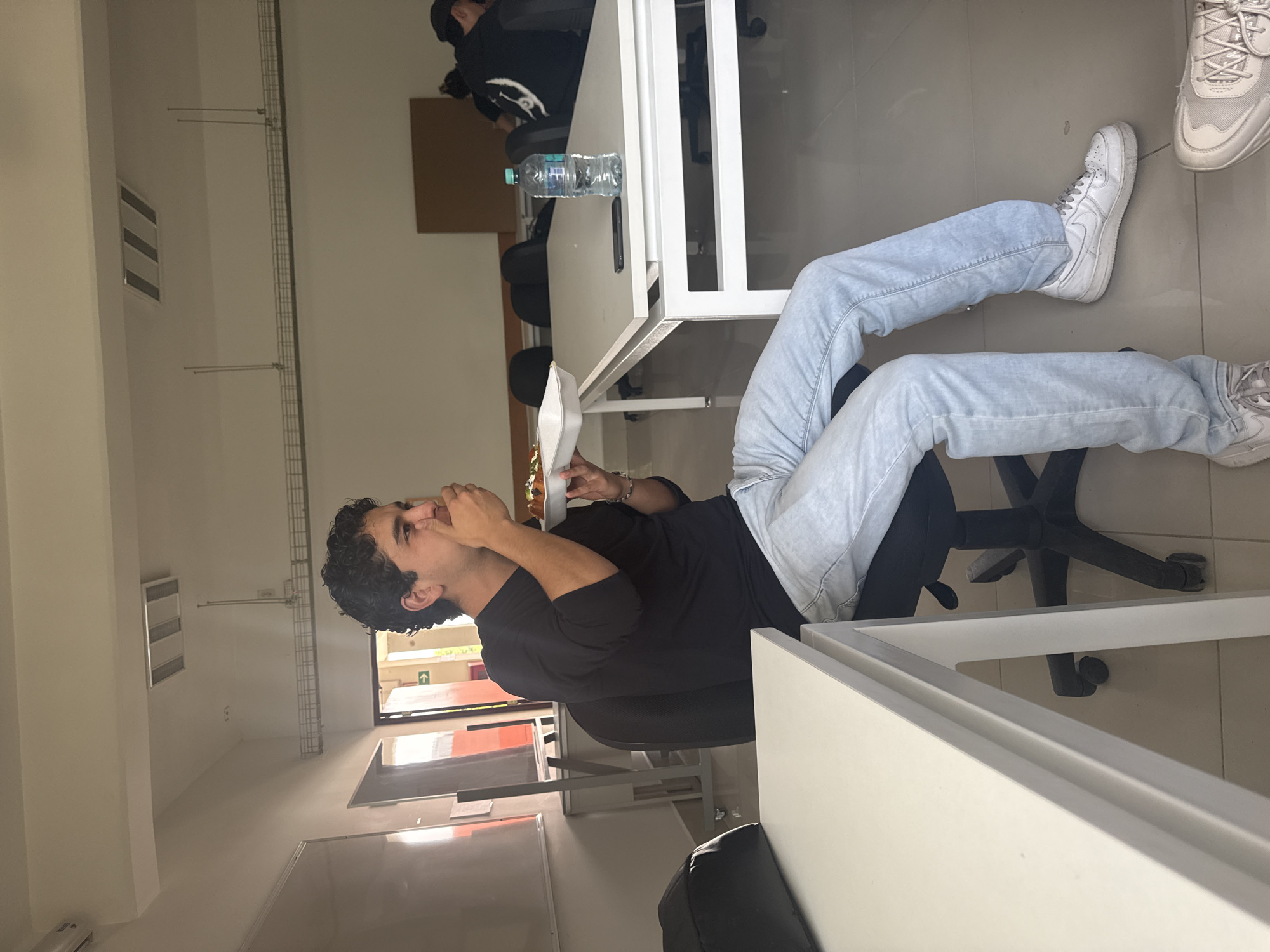 A man is sitting in an office chair, positioned with his legs stretched and elevated onto a white table. He is wearing light blue jeans, a black shirt, and white sneakers. In his hand, he is holding a white container, possibly a food container, near his face. The office space contains white tables, other chairs, and a person working in the background. A water bottle is placed on one of the tables. The setting appears to be a modern office environment. The location is San Andrés Cholula, Mexico.