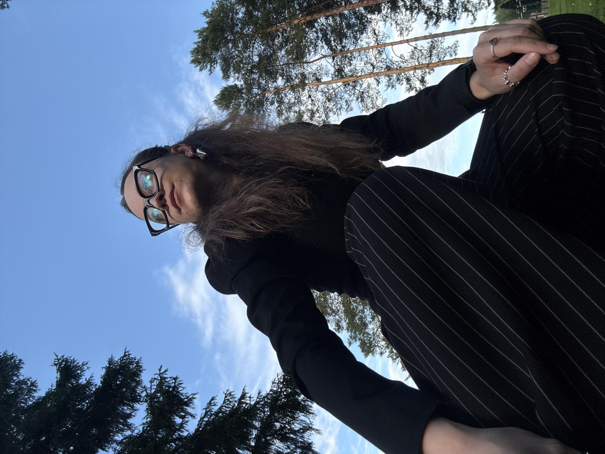 A person wearing a black blazer and pinstripe pants crouches outdoors under a partly cloudy sky. The person has long, dark, wavy hair, is wearing black-rimmed glasses, and has a silver heart-shaped earring in their left ear. They are looking upwards and to their left. Their right hand is resting on their right knee, displaying two rings. The background features several tall pine trees and a glimpse of a green grassy area, possibly a park or the outskirts of a town. No specific landmarks are visible to identify the location as Votkinsk, Russia.