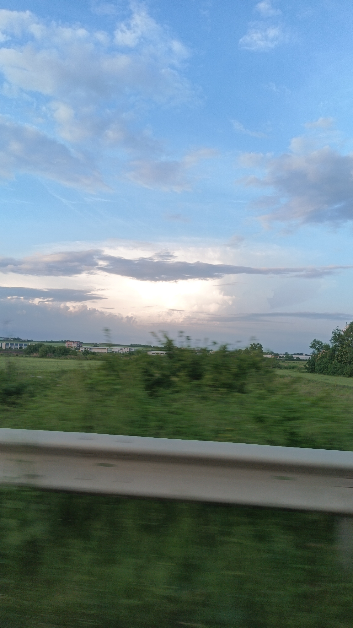 The image presents a view from a moving vehicle, likely on a highway. The foreground is a blurred view of green vegetation and a roadside barrier. Beyond this, a green field stretches toward the horizon. In the distance, there are structures that appear to be buildings or industrial facilities. The sky is partially cloudy with blue patches. Based on the landscape and buildings in the background, the location could potentially be near Burgas, Bulgaria, although specific architectural or geographical features confirming this are not discernable in this shot. The absence of distinct people and objects in the foreground contributes to a general impression of movement through a rural setting.