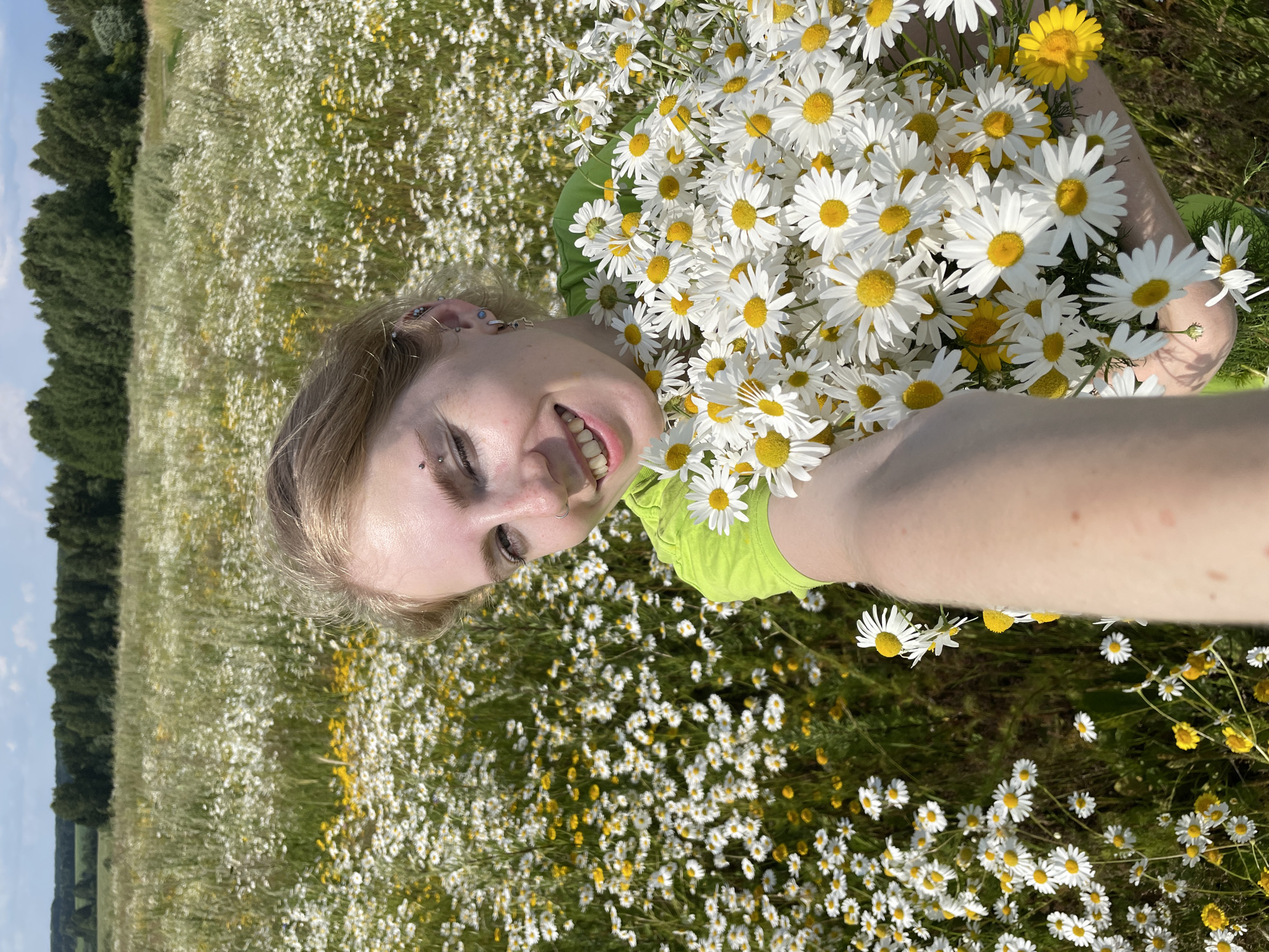 A person is lying in a field of daisies, holding a large bouquet of the flowers. The person is wearing a bright green shirt and has blonde hair. They are smiling and looking at the camera. The field is filled with white and yellow daisies, with a line of trees in the background. The sky is blue and there are some clouds. The time of day appears to be daytime, with bright sunlight. The location is identified as Сям-Можга, Russia. The overall mood is joyful and peaceful. There is no visible text in the image.