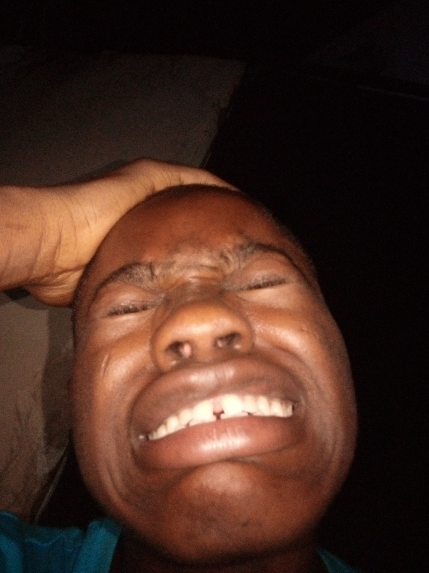 A close-up, low-angle, full-face shot captures a Black individual with their eyes closed and mouth open in a grimace. A hand is visible at the top of the frame, resting on the individual's head. The background is dark and indistinct, suggesting an indoor setting at night. The individual is wearing a teal-colored garment. The image appears to be taken in Shangisha, Nigeria.
