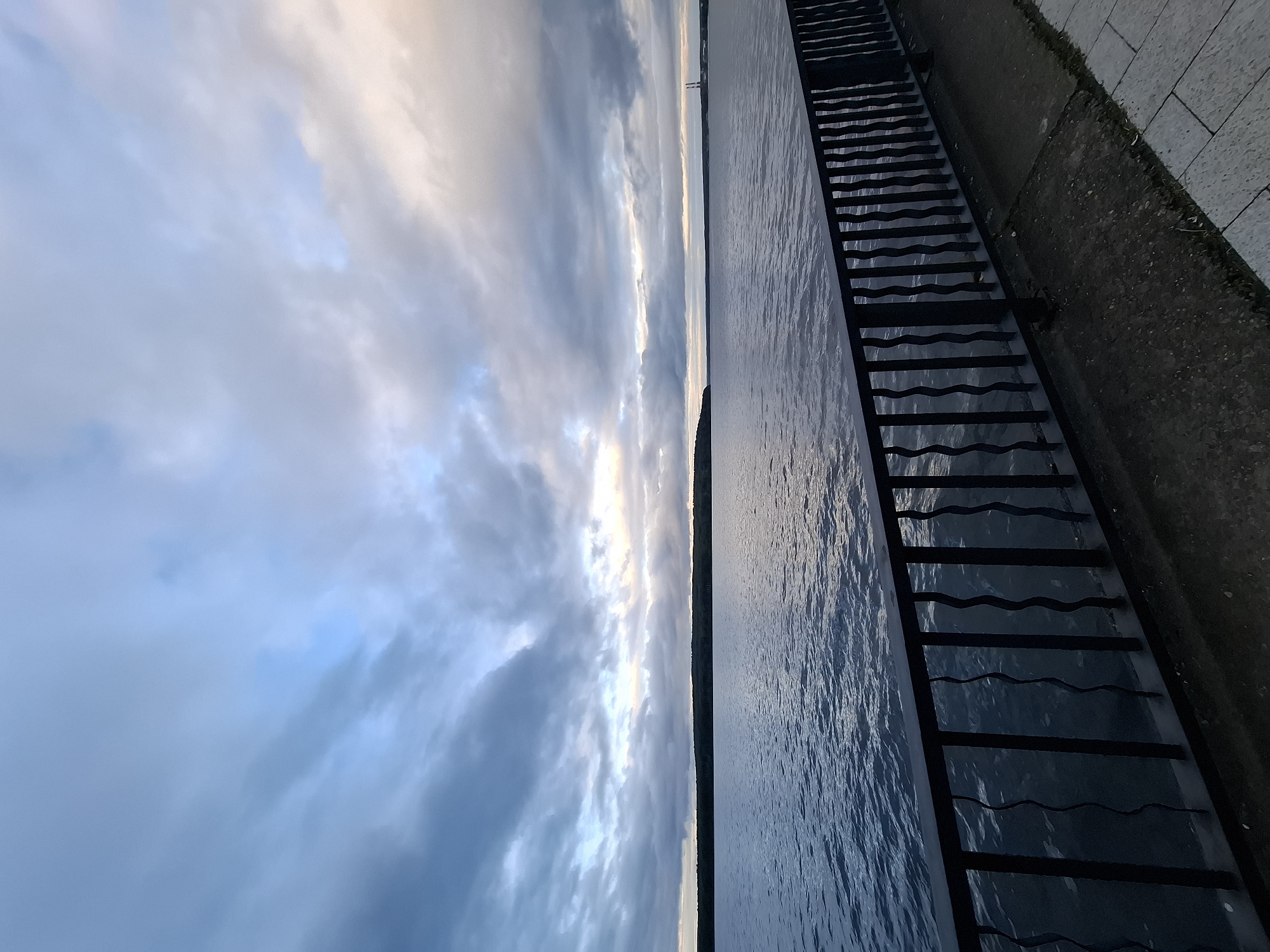 The image depicts a panoramic view of a large body of water, likely the Votkinsk Reservoir on the Kama River, bordering Chaikovsky, Russia, at twilight. The sky is extensively covered by thick, layered gray and blue clouds, with faint streaks of pale orange and white light visible towards the distant horizon. The wide expanse of water below is dark blue-gray with subtle surface ripples, reflecting the subdued light from the sky. A continuous, dark horizontal line, characteristic of a forested landmass or distant bank, separates the water from the far horizon. On the distant right horizon, two tall, slender industrial structures are discernible against the lighter sky. In the foreground, a dark metal fence with distinctive wavy vertical bars extends horizontally across the frame, positioned above a concrete embankment. The ground directly beneath the fence is dark, textured concrete, transitioning to lighter, rectangular paving stones on the right side of the frame. No individuals or active subjects are present in the scene.