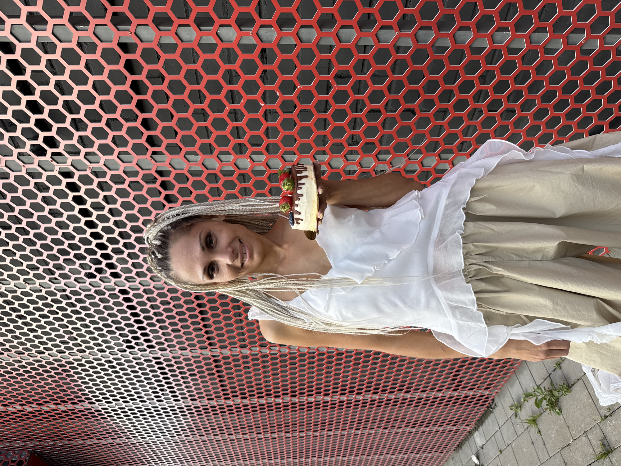 A woman with blonde braided hair stands in front of a red honeycomb-patterned wall, holding a small cake topped with strawberries and blueberries. She is wearing a white ruffled top and beige pants. The background appears to be an outdoor urban setting, with a cobblestone path and some greenery visible on the ground. The image was taken in Есбол ауылдық округі, Kazakhstan.