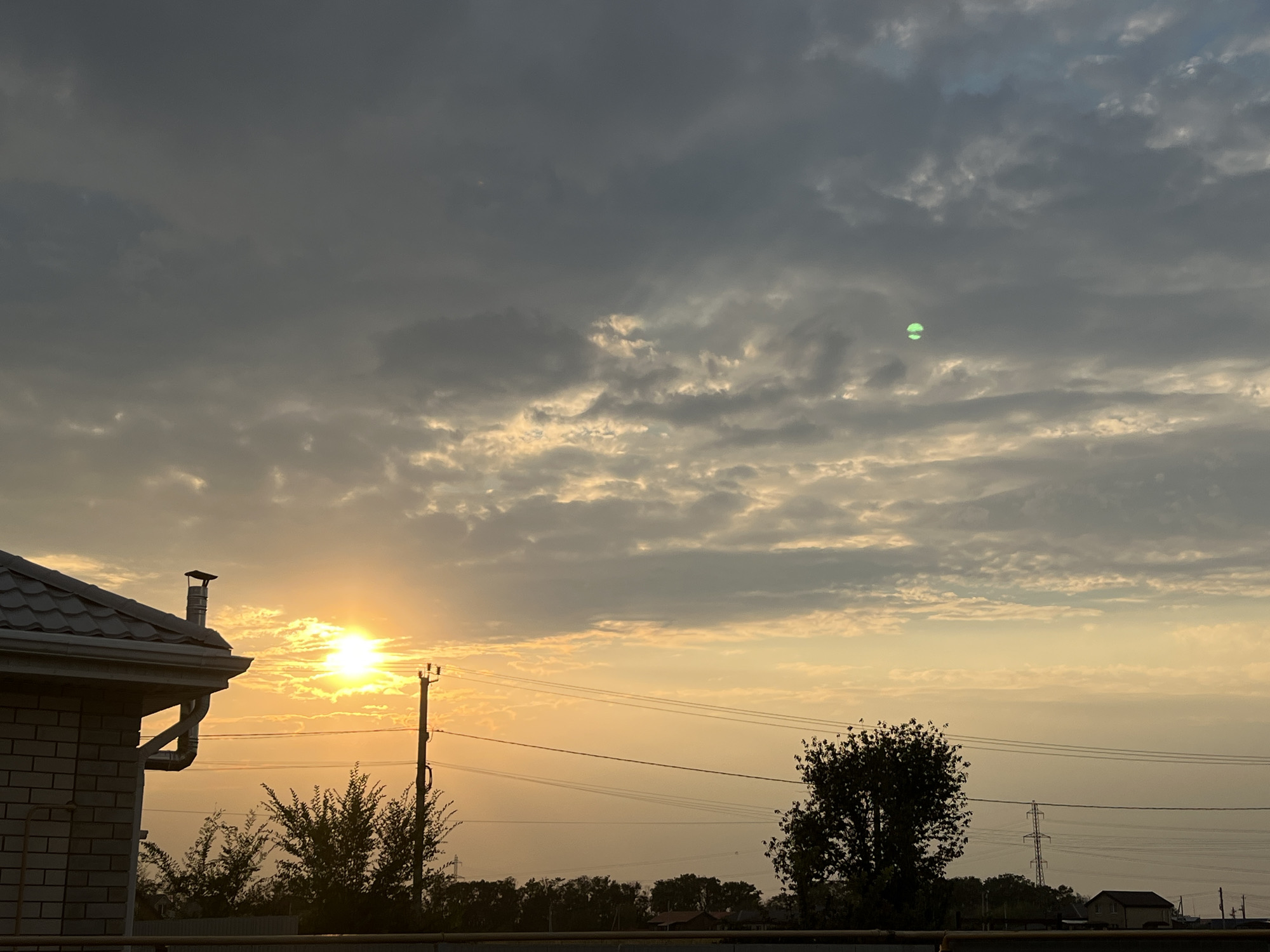 The image captures a sunset scene in the city of Taganrog City District, Russia. The sky is filled with dramatic clouds, tinged with orange and yellow hues from the setting sun, which is partially obscured by a band of clouds on the left side of the frame. A bright, luminous sphere, appearing to be a green light source, is visible in the upper right quadrant of the sky. In the foreground, the roof and part of a brick building are visible on the left, with a chimney pipe extending from the roof. A telephone pole with wires stretches across the middle ground, and silhouetted trees are present beneath the horizon. The overall environment appears to be a semi-rural or suburban area.