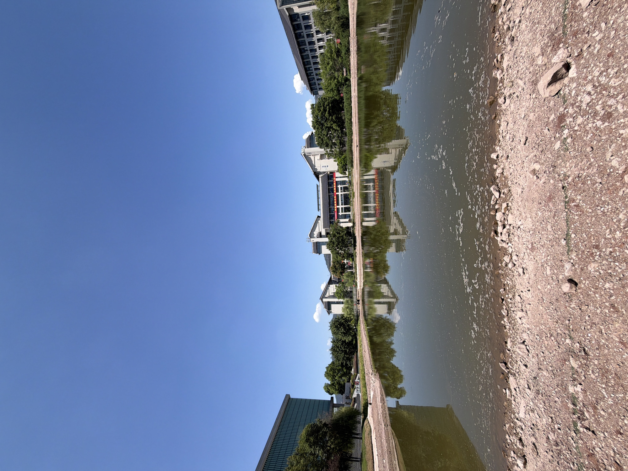 In the city of Jiangjin, China, a wide-angle, eye-level shot captures a serene scene featuring a calm lake reflecting buildings and trees under a clear blue sky. The foreground is dominated by a rocky, reddish-brown shore, scattered with pebbles and larger stones. The lake's surface, with a slightly murky green hue, exhibits a gentle ripple and a scattering of white foam or debris.
Across the water, a line of modern buildings with dark roofs and light-colored facades stretches horizontally. The buildings vary in height and architectural style, some featuring multiple stories and numerous windows. Lush green trees are interspersed between the structures, adding a natural element to the urban landscape. Red banners with white Chinese characters are visible on some of the buildings, suggesting institutional or governmental occupancy. The reflection of the buildings and trees is clearly visible on the still surface of the lake, creating a symmetrical visual effect. The sky above is a vibrant, cloudless blue. There are no people or animals visible in the image.