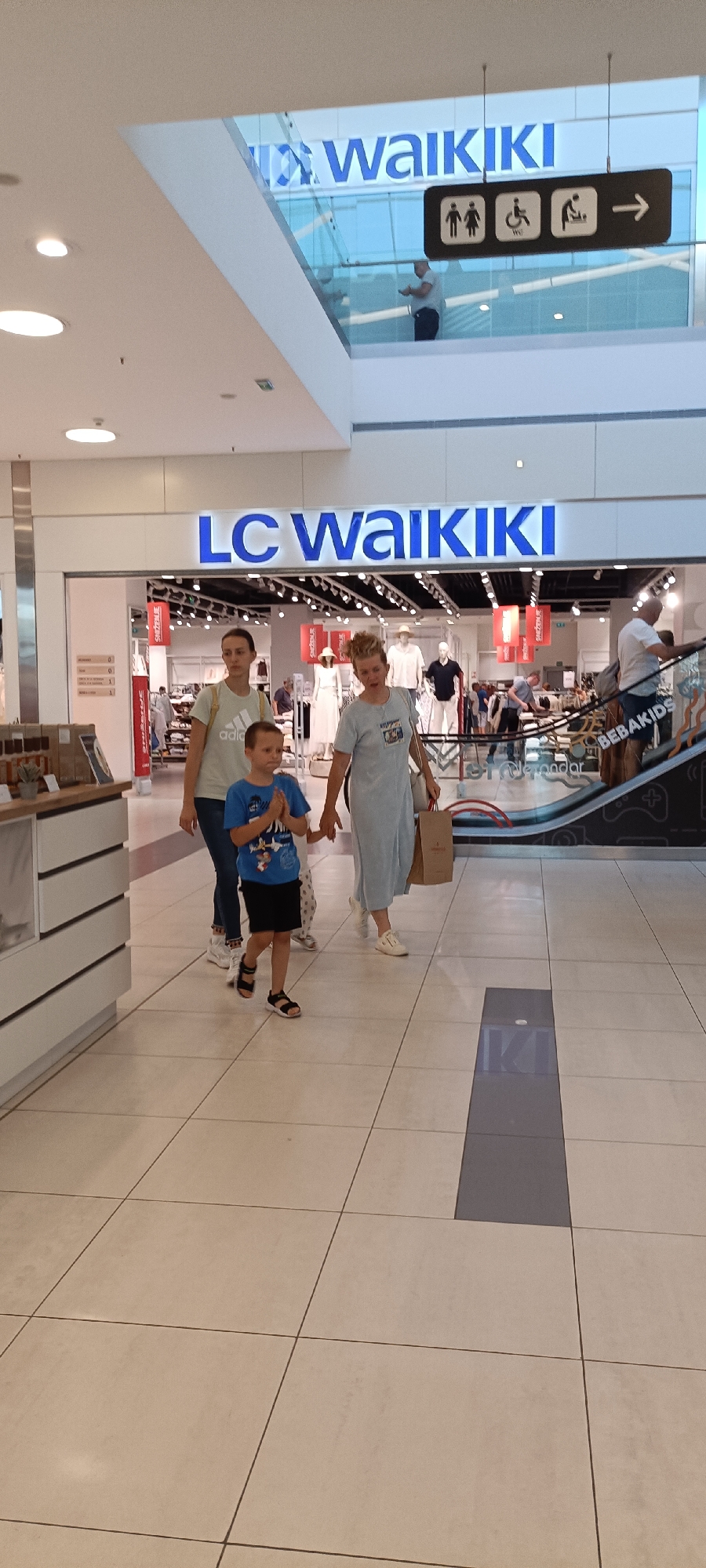 This is a vertical, eye-level, full-shot photograph taken inside a brightly lit, modern shopping mall. The main subjects are three people walking from left to right: a woman in a light green t-shirt, dark pants, and white sneakers, holding a brown paper shopping bag; a young boy in a blue t-shirt and black shorts with black sandals, his hands clasped in front of him; and another woman in a light blue dress and white sandals, also carrying a brown paper shopping bag. The boy appears to be looking towards his right, while the women are looking forward.
Behind them, the entrance to a store is visible with a prominent blue sign that reads "LC Waikiki". Mannequins displaying clothing are visible inside the store. Further in the background, an escalator is ascending, with people using it. To the right of the escalator, another sign reads "BEBAKIDS".
Above and behind the entrance to LC Waikiki, a sign for "WAIKIKI" is visible in large blue letters. To the right of this sign, a dark rectangular sign with white icons depicts figures of a man, a woman, a person in a wheelchair, and a baby changing station, with an arrow pointing to the right. A man in a light blue shirt and dark pants is visible standing on a balcony above the store entrance, looking at his phone.
The floor is tiled with large, light-colored square tiles, with a darker, rectangular inlay running down the center of the walkway. The overall impression is of a typical indoor shopping environment during daylight hours, with people engaged in shopping activities. The image appears to have been taken in Novi Sad, Serbia, given the presence of a "LC Waikiki" store, a popular clothing retailer in the region.