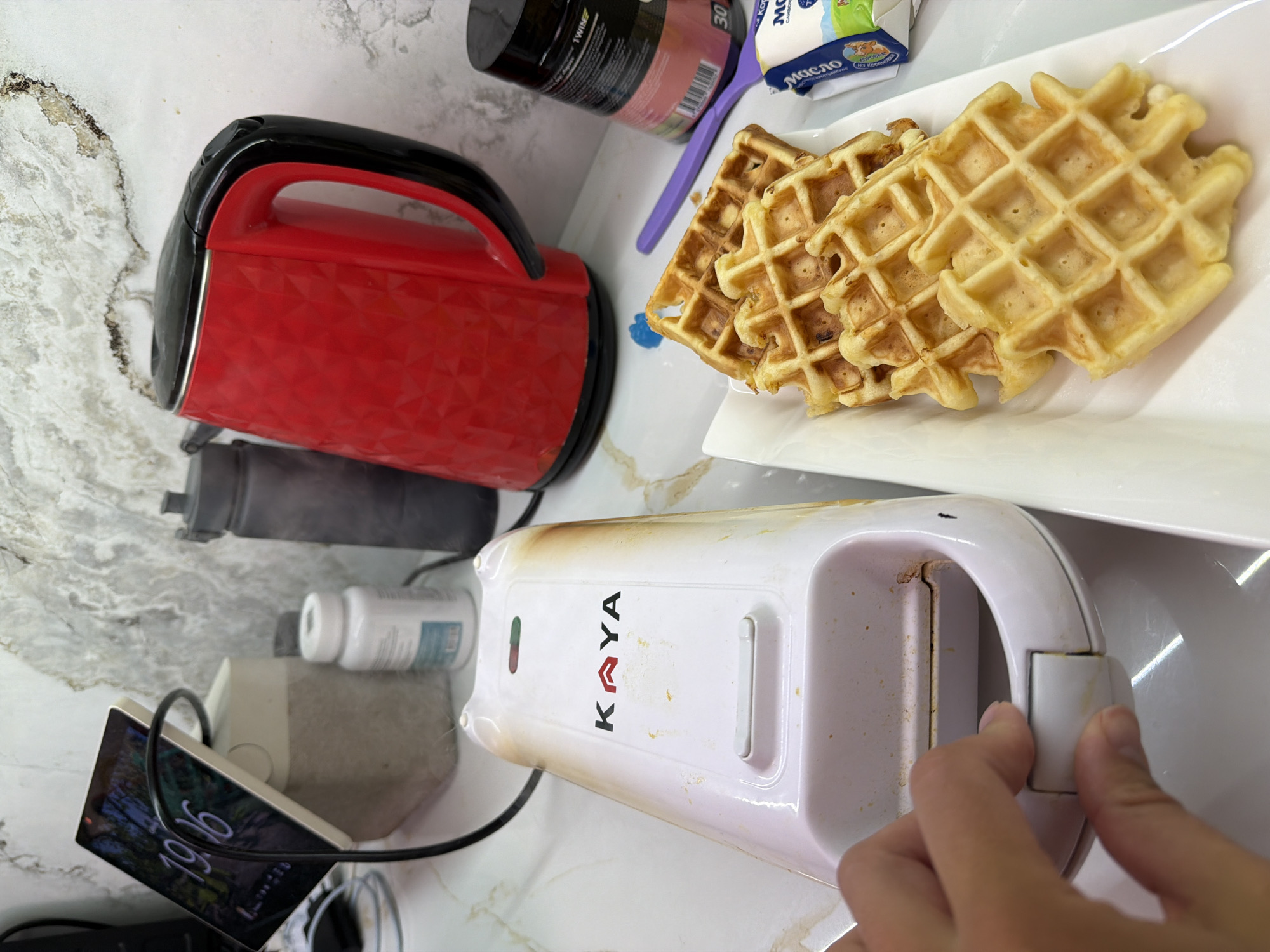 The scene is a countertop, possibly in a kitchen. Present are waffles on a white plate, a red waffle maker, and a white waffle maker with the brand name "KAYA". Also visible are a black supplement container, a container of butter, a purple spoon, a water bottle, a pill bottle, a phone, and other unidentifiable objects. A hand is seen operating the white waffle maker. There is no visible evidence to suggest that this image was taken in Sochi, Russia.