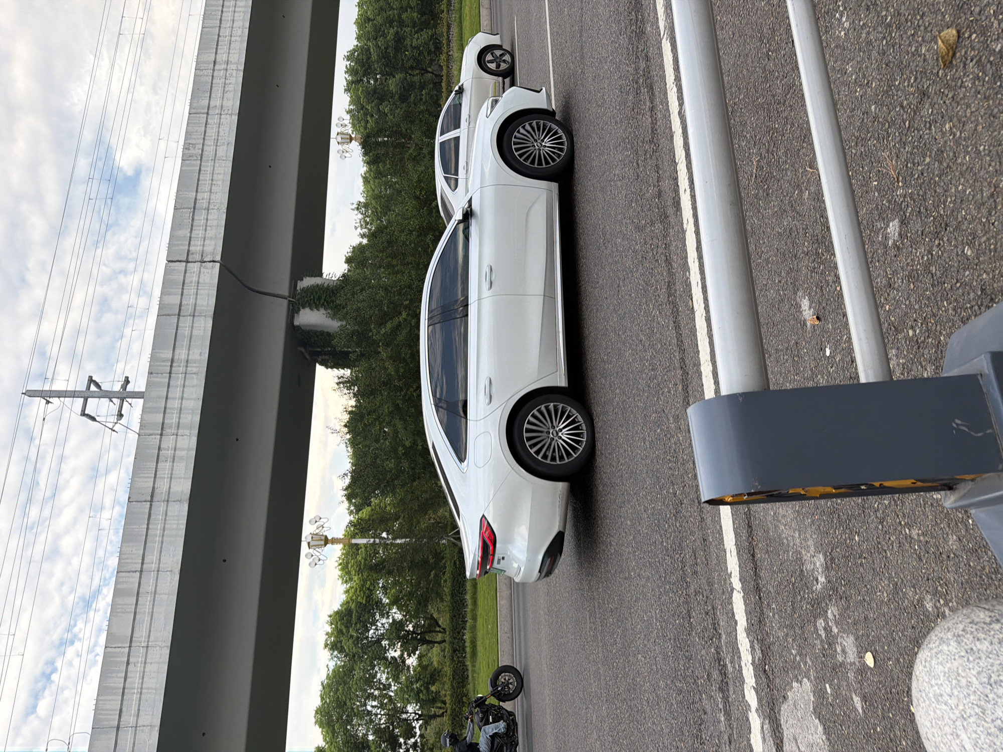 The image captures a street scene in Jiangjin, China, characterized by an elevated road or bridge structure dominating the upper portion of the frame. Below this structure, the main road is visible, with two white cars driving. The car closer to the foreground is a modern sedan with distinctive taillights and alloy wheels. Further down the road, another white car is visible. To the left, a person is riding a motorcycle, also on the road. The environment includes a line of trees and greenery along the side of the road, and a cloudy sky. In the foreground, a gray protective barrier or bollard is partially visible. The overall impression is of a typical urban or suburban transit route under an overpass.