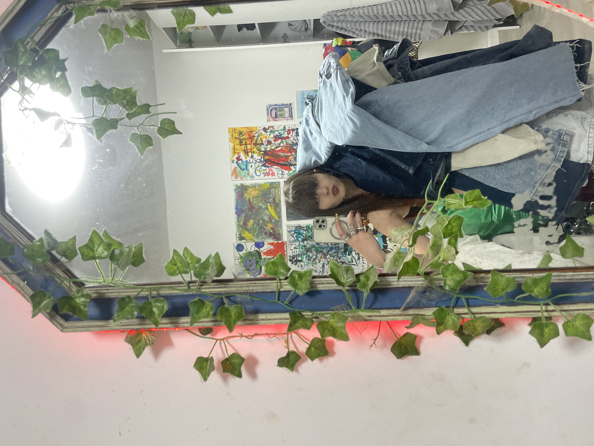 A selfie is taken of a person in the mirror in a room. The mirror frame is adorned with fake green ivy and red LED lights. In the mirror's reflection, the person has dark hair and bangs. They are holding a phone with two rear cameras. Behind the person in the mirror's reflection, there are paintings on the wall. The location is unknown so I cannot tell you if it is in the city of Raanana, Israel. A pile of clothes are stacked. It's impossible to determine the location and specific details about the room or any actions or interactions, as only a reflection in the mirror is visible.