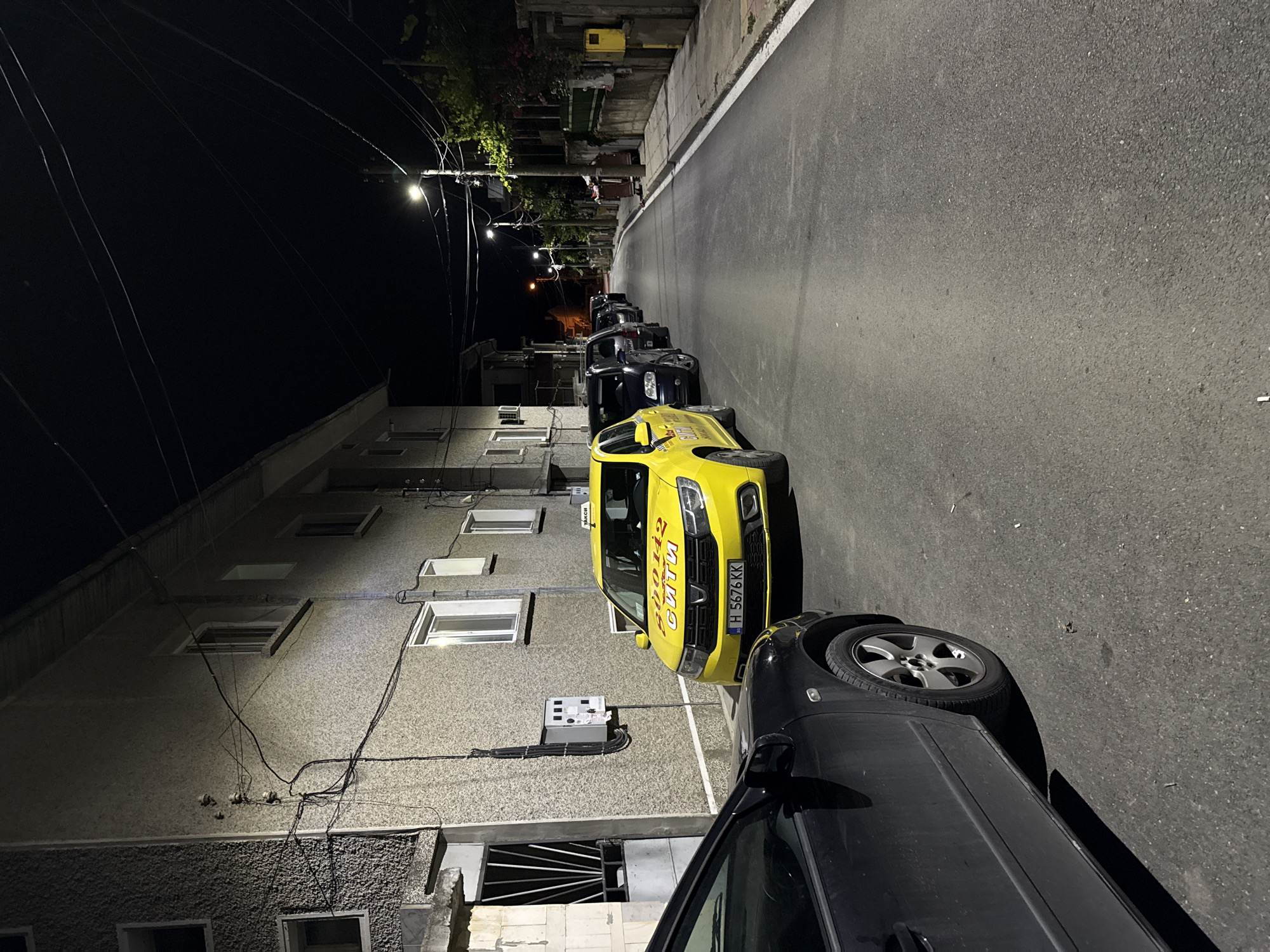 The image shows a street scene at night in Shumen, Bulgaria. A yellow taxi with the number plate H 5676 KK is parked on the side of the road, with a black car in the foreground. Other cars are parked further down the street. The buildings on the left appear to be residential, with windows and electrical wiring visible. Streetlights illuminate the scene, casting shadows on the asphalt road. The environment is urban, with sidewalks, street signs, and a few trash bins visible in the distance. The overall atmosphere is quiet and still, typical of a residential street at night.