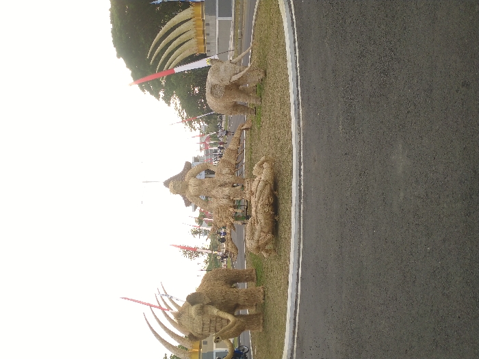 The image depicts a daytime scene at a public roundabout or large circular median in Sragen Kulon, Indonesia. The central grassy area of the median features a collection of large sculptures constructed from light-brown straw. A prominent humanoid figure, approximately 3-4 meters in height, stands centrally, positioned above several smaller, recumbent straw forms on the ground. To the left, a large straw sculpture resembling a woolly mammoth with long, curved tusks is present. On the right, another large straw sculpture, appearing as an elephant or mammoth with a lowered trunk, faces left. The immediate foreground consists of a dark asphalt road surface. In the background, an urban environment is visible, characterized by numerous red and white flags mounted on poles and interspersed with green trees. A substantial monument or structure, composed of multiple upward-curving, tusk-like forms, is situated on the far right. Several human figures are observable in the distant background, with one partially visible individual on a motorcycle on the far left. The sky is uniformly bright and clear, indicating daylight conditions.