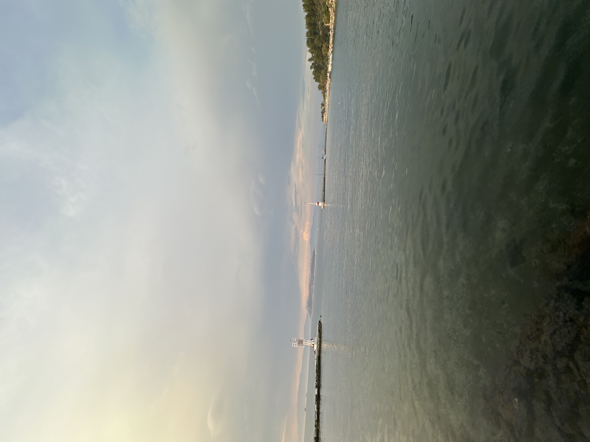 The image captures a serene coastal scene in the city of Thassos, Greece, likely at sunset or under overcast skies. In the foreground, the calm, dark water of the Aegean Sea fills the lower half of the frame. The surface of the water shows gentle ripples, with some submerged rocks visible near the bottom left.
The mid-ground is dominated by the expansive sea and the distant shoreline. To the left, a breakwater made of large stones extends into the water, topped by a distinctive white and red lighthouse. This structure appears to be a navigational aid for the harbor. Further along the breakwater, a few small, dark shapes suggest boats or other harbor features.
In the distance, the coastline of Thassos is visible, characterized by rolling hills covered with lush green trees. A second, smaller lighthouse or navigational marker stands on a point of land further along the coast, closer to the right side of the image. The sky above is a mix of soft, pale yellow and blue hues, with wispy clouds indicating changing weather conditions, possibly the remnants of a recent rain or the approach of evening. The overall atmosphere is tranquil and picturesque, highlighting the natural beauty of the Greek island.