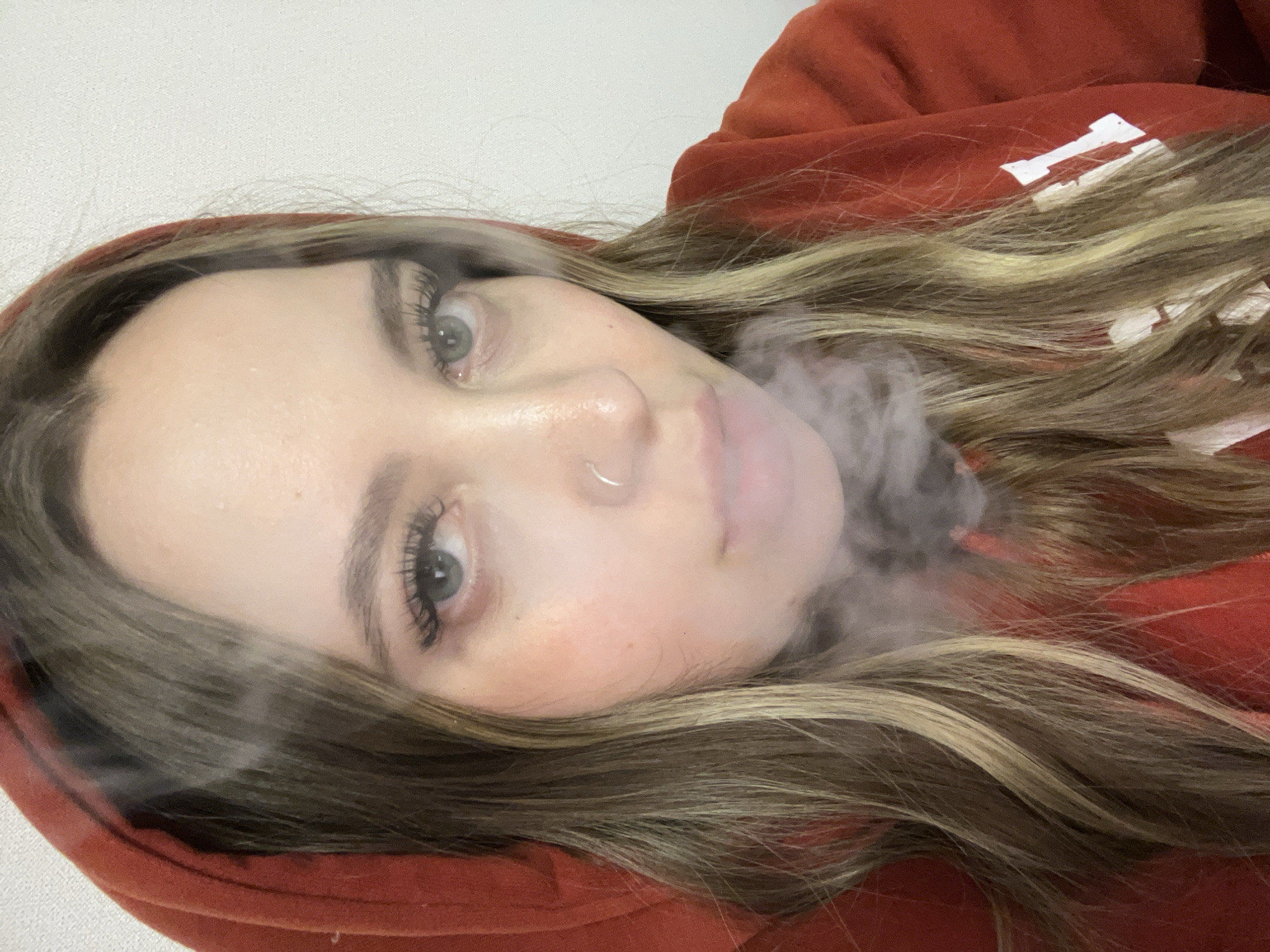 A young woman with long blonde hair wearing a red hooded sweatshirt is exhaling a cloud of smoke. She has a nose ring and is looking directly at the camera. The background is a plain white wall. No information indicates that the location is Krasnodar, Russia.