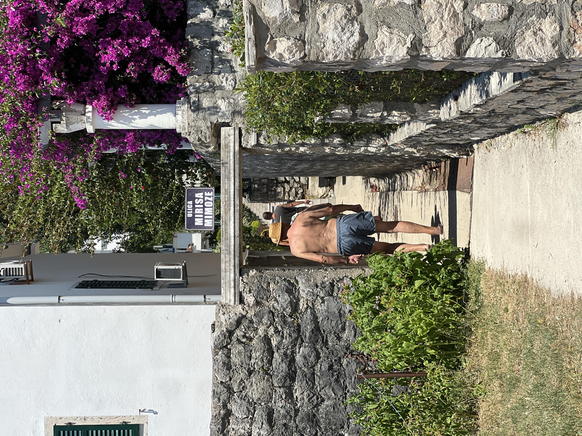 A shirtless man wearing a straw hat and blue shorts walks along a paved path next to a stone wall. He is facing away from the camera, with his right hand resting on his hip and his left arm slightly bent. The path appears to be in an outdoor setting, possibly a coastal town given the presence of lush green foliage and flowering plants. To the left of the path, a white building with a dark green window shutter is visible. A sign with black lettering on a white background reads "ULICA MIRISA MIMOZE". The time of day appears to be sunny, with distinct shadows cast on the ground. The overall scene evokes a relaxed, summery atmosphere.