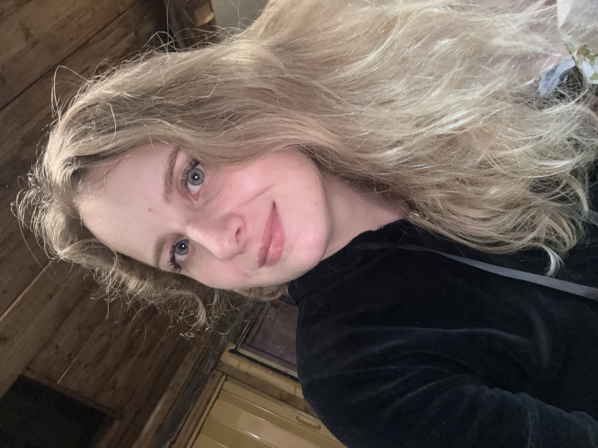The image is a close-up, indoor selfie of a young Caucasian woman with blonde, wavy hair and blue eyes. She is wearing a black hooded jacket. The background consists of wooden paneling on the ceiling and walls, suggesting a rustic or cabin-like environment. The lighting is warm and appears to be artificial. There are no other people or significant objects visible in the frame. The image does not provide any specific location details within Severnyy, Russia.