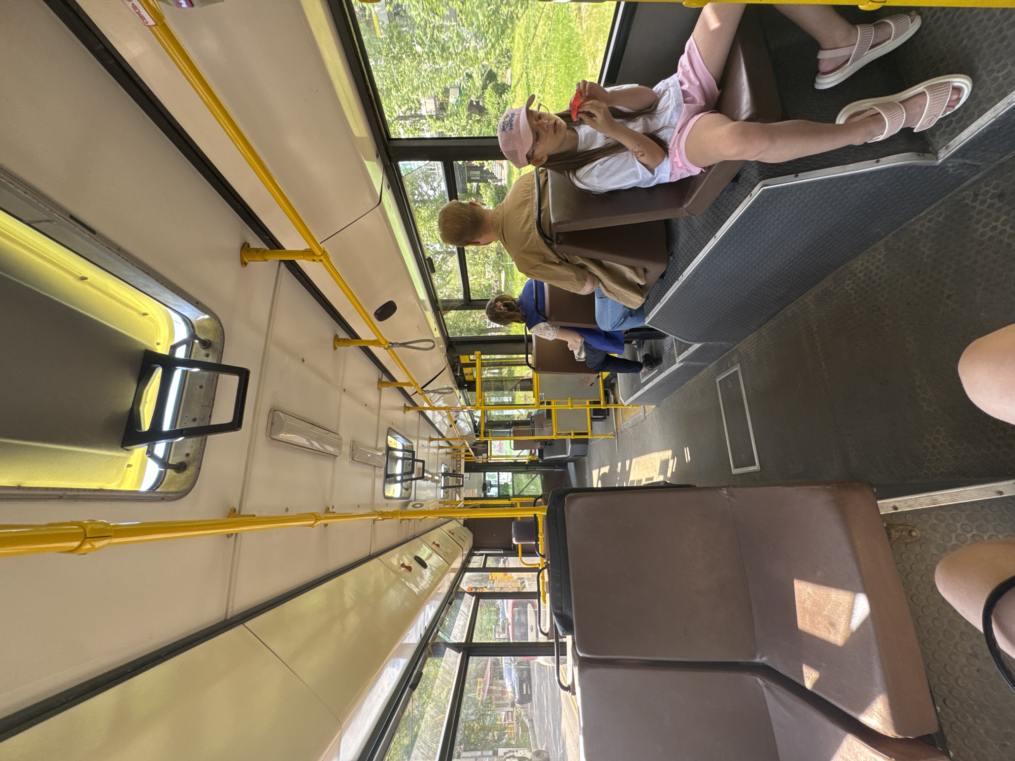 The interior of a public bus is depicted, focusing on the seating arrangement and passengers. A girl with a pink cap, white shirt, and pink shorts is seated near the window, engaged with a red object. An adult with a light brown jacket sits nearby. Further back, a woman with a blue dress is partially visible. The bus has yellow handrails and windows revealing greenery outside, suggesting a ride through a suburban or semi-rural area. The seats are brown, and the floor is dark gray. There are more seats towards the front. The location is in the city of Unknown, Unknown.
