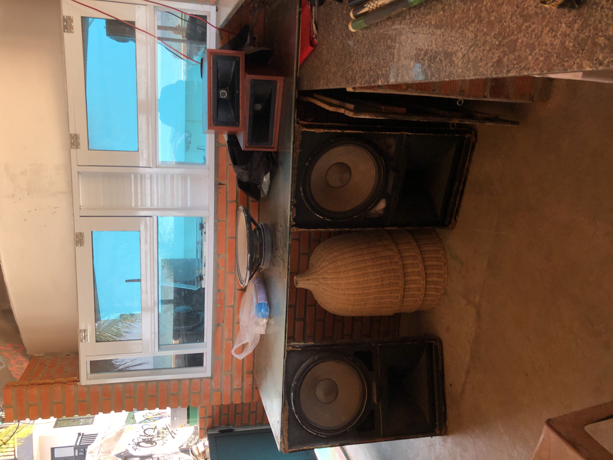 The image depicts a room with brick walls and a large window overlooking a body of water, likely the ocean. The room is filled with audio equipment, including large speakers, smaller horn speakers, and what appears to be a speaker cone. A wicker basket-like object is stacked on the floor. The scene suggests a workspace or storage area for music equipment, possibly in a coastal town like Mũi Né, Vietnam, given the visual cues of the water and architectural style. The overall impression is one of functional clutter.