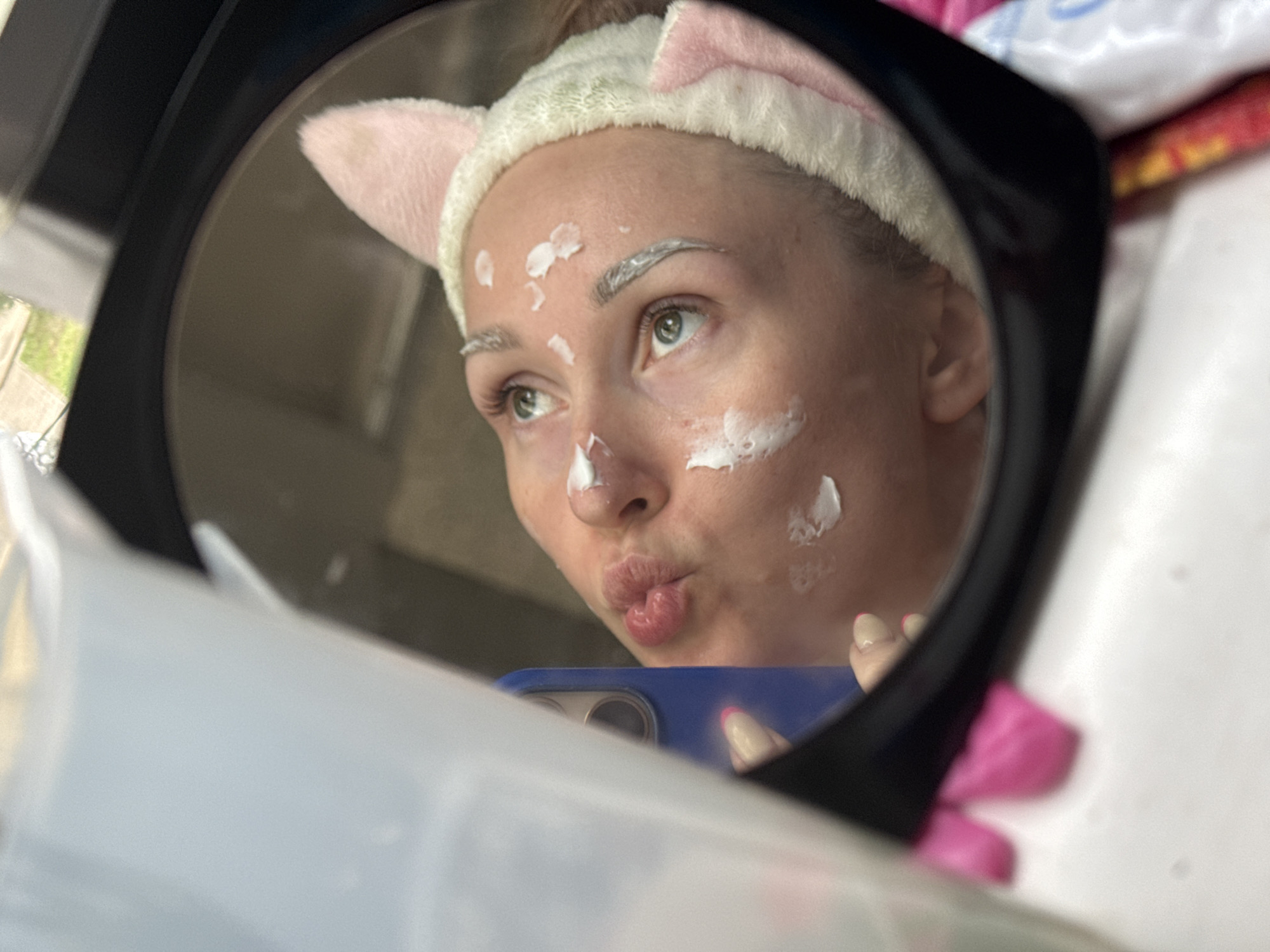 A woman wearing a cat-ear headband is reflected in a black-framed mirror. She has white cream or lotion applied to her face and eyebrows. She is holding a blue smartphone, taking a selfie and making a kissing face. The immediate environment around the mirror includes miscellaneous objects with pink and white colors. The specific location is unknown.