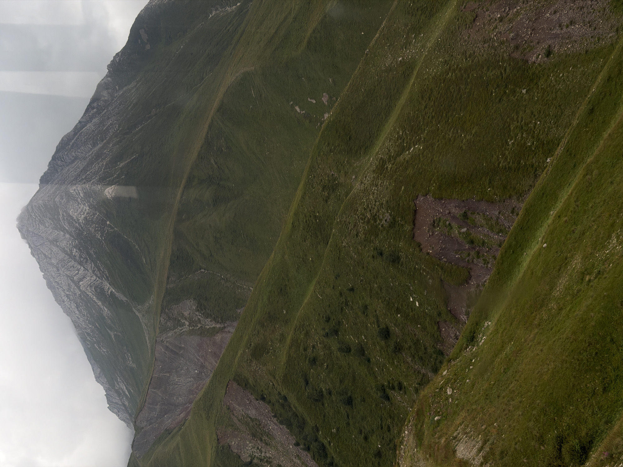 The image shows a view of a mountain range covered in green grass and rocky slopes. The sky is overcast, and the lighting is subdued, suggesting it might be a cloudy or foggy day. There are no visible people, structures, or specific landmarks that would indicate a location within the city of Almasiani, Georgia. The scene is a natural, mountainous landscape.