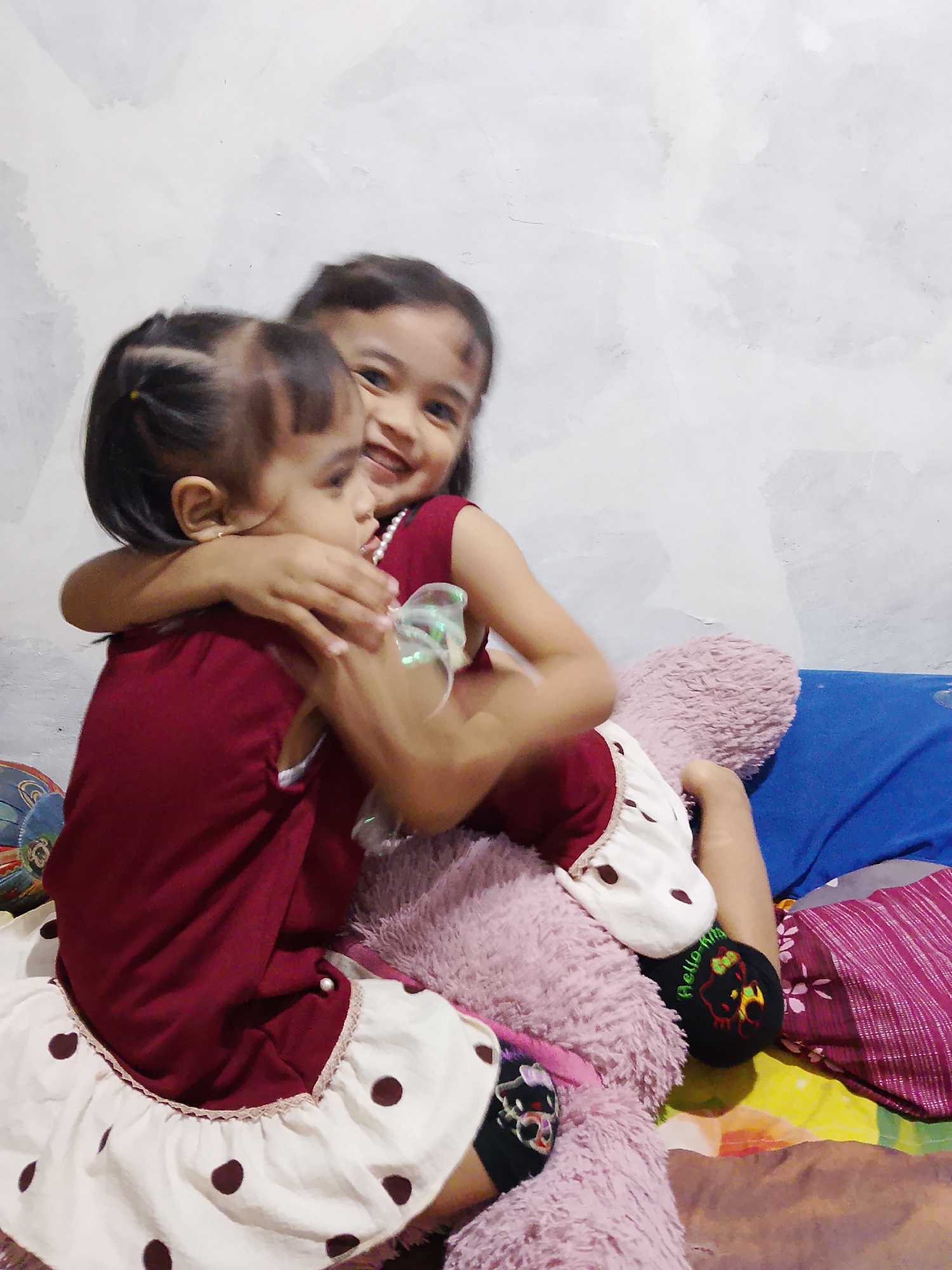 This media file captures a candid moment between two young girls. The girl in front, facing away from the camera, is wearing a dark red sleeveless top and a white skirt adorned with dark brown polka dots. She has her hair styled in two pigtails with yellow ties. The girl behind her, who is facing the camera, is also wearing a dark red sleeveless top and appears to be wearing a white necklace. She has a wide, joyful smile. The girls are embracing each other tightly, with the older girl's arms wrapped around the younger one. They are seated on a surface that appears to be a bed, with a large, fluffy pink stuffed animal between them. Other objects visible include a blue pillow on the right and a patterned blanket or cloth in the background on the right. One of the girls is wearing a black sock with an embroidered image of Hello Kitty and the text "Hello-Ki". The setting seems to be indoors, likely a bedroom, with a textured light-colored wall behind them. The lighting suggests it is daytime, but there are no specific indicators of the time of day or weather. The overall emotion conveyed is one of affection and happiness between the sisters. The location context is Tanggungan, Indonesia.
