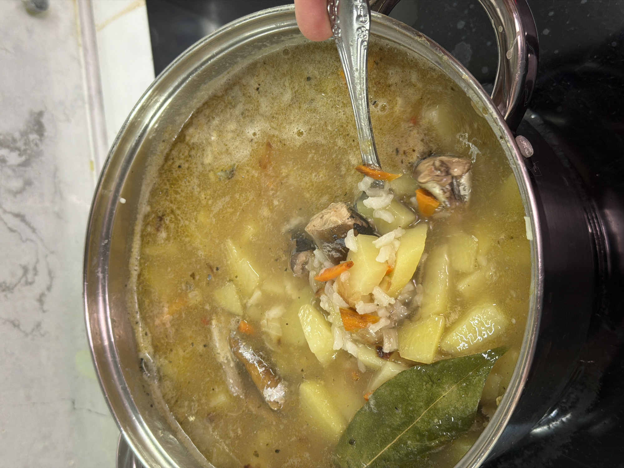 A close-up, overhead shot shows a pot of fish soup with potatoes, rice, carrots, and bay leaves. A spoon is scooping some of the soup out of the pot. The soup is a light yellow color and appears to be steaming. The pot is made of stainless steel and is sitting on a black stovetop. The background is a blurry image of a marble countertop. There is no indication of the location in Sochi, Russia.