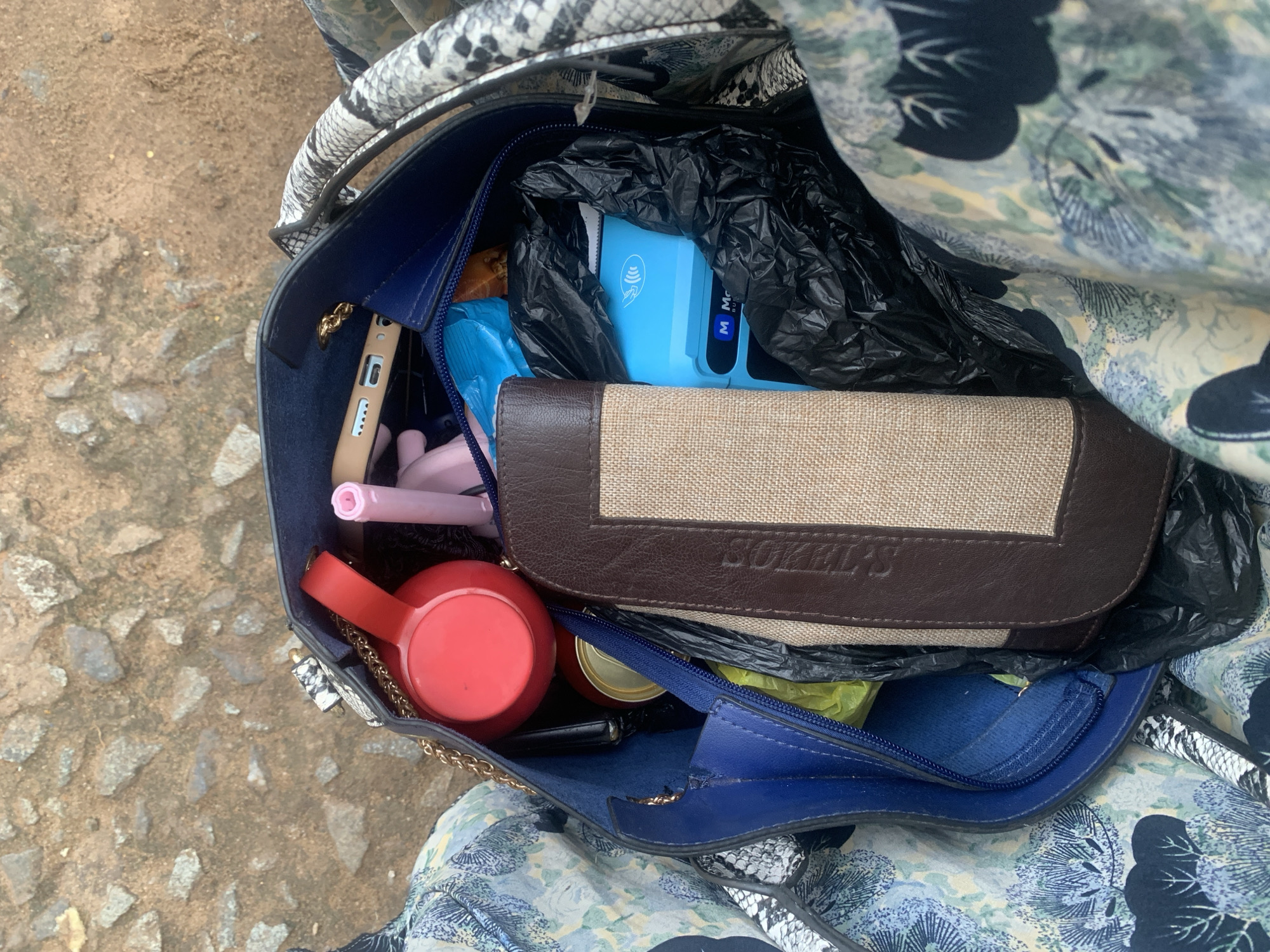 A high-angle, full shot captures the interior of a dark blue tote bag with a snakeskin patterned handle, resting on a textured, rocky ground. The bag is filled with various items, including a red circular object, a pink makeup item, a light brown wallet with a textured beige insert, a blue card reader, and a can. The ground appears to be dirt and gravel, consistent with an outdoor urban environment. The overall scene suggests a snapshot of everyday items within a personal bag in Elelenwo, Nigeria.