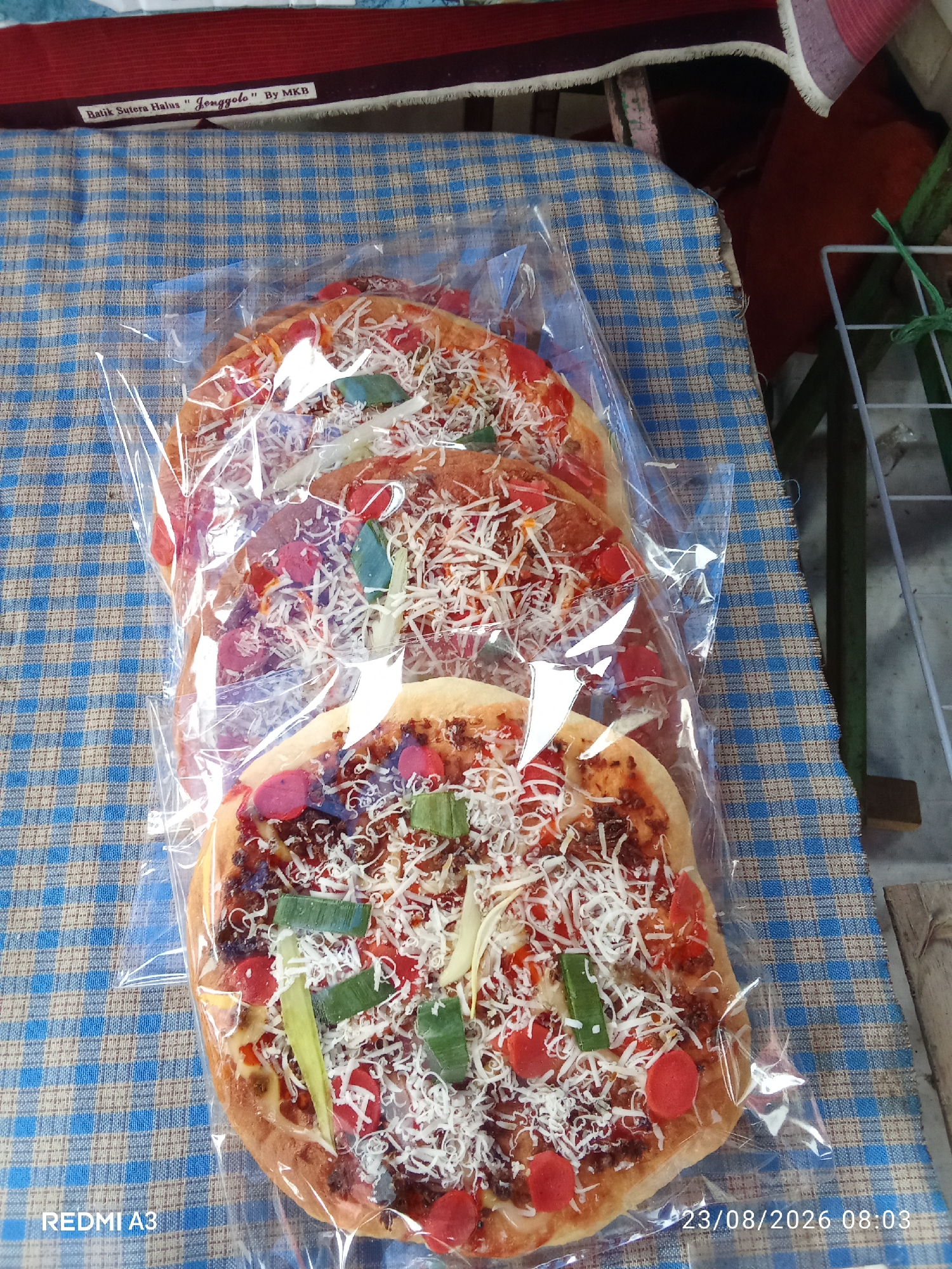 The image depicts three small, individually packaged pizzas arranged on a blue and tan checkered tablecloth. The pizzas appear to be topped with shredded cheese, tomato sauce, possibly some meat, and green bell pepper pieces. One pizza is slightly more in focus, showing sliced red sausage-like pieces as well. The tablecloth is laid out on a flat surface, and a portion of a dark red and burgundy striped fabric with a white fringe is visible at the top. A white tag attached to the striped fabric reads "Balik Sutera Halus "Jenggolo" By MKB". A metal rack is visible in the background to the right. The bottom left corner of the image has a watermark that reads "REDMI A3", and the bottom right corner has a timestamp: "23/08/2026 08:03". The scene is indoors or in a covered outdoor space, likely a market stall or shop given the presentation of goods.