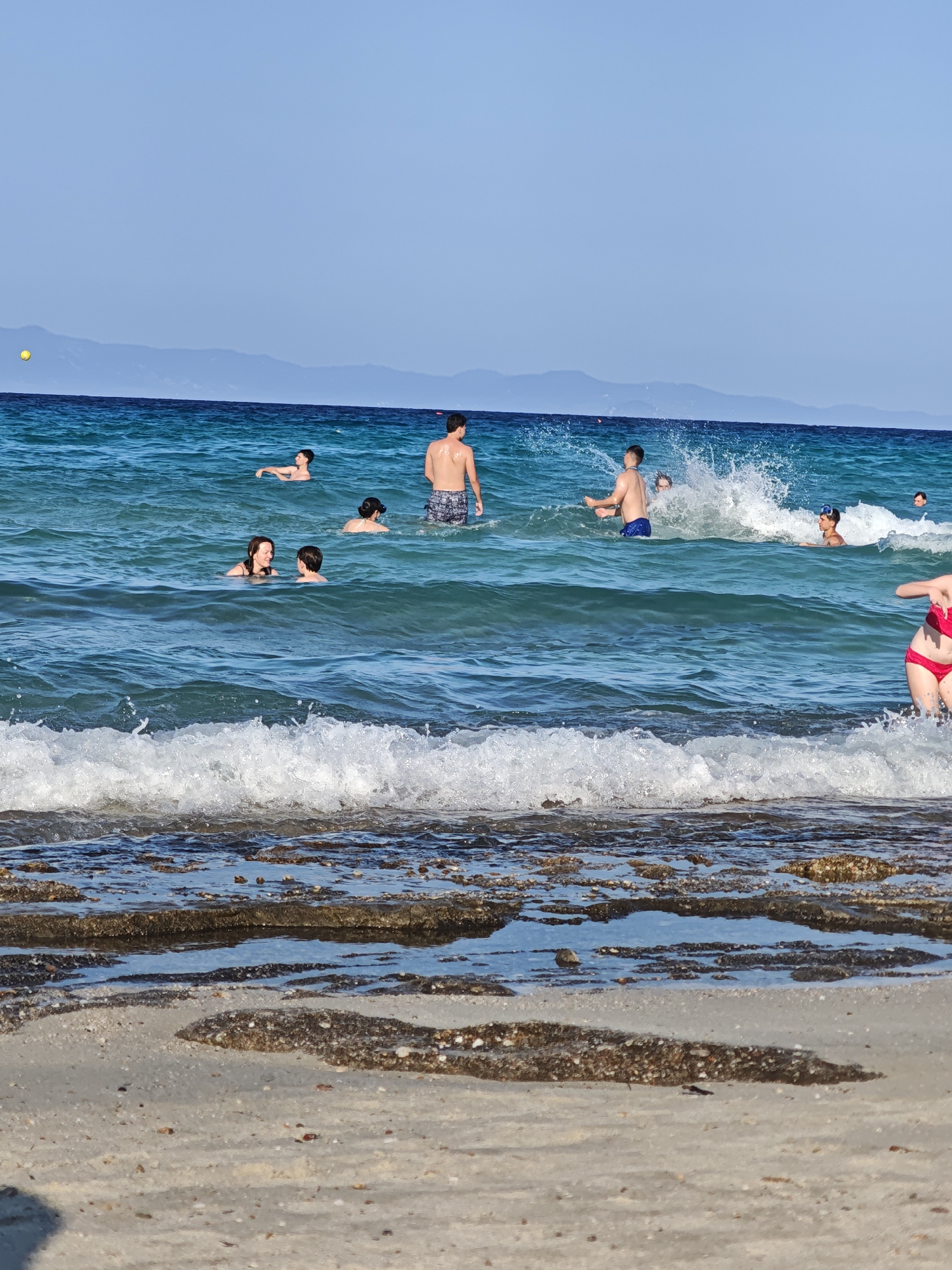 In the city of Kassandra Municipal Unit, Greece, a group of people are enjoying a sunny day at the beach. The image captures a view from the shore, looking out onto the sea. The water is a vibrant blue, with gentle waves breaking near the beach. Several individuals are visible in the water, swimming and playing. In the foreground, the sandy beach is visible, with some rocky formations partially submerged in the shallow water. The sky above is clear and blue, with distant mountains forming the horizon line. The overall scene depicts a relaxed and enjoyable beach atmosphere.