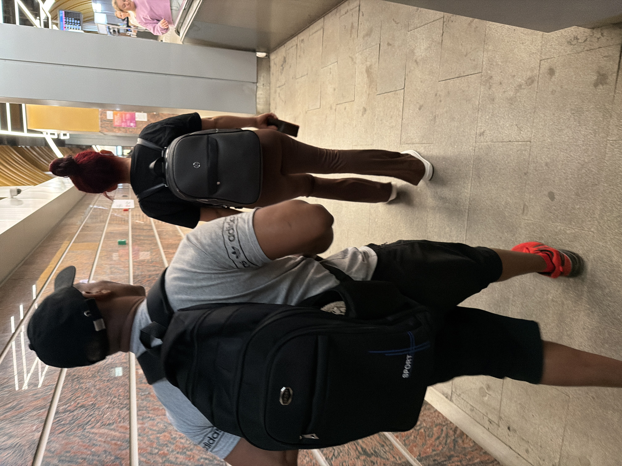 The image shows two people walking down a tiled hallway, likely in a Moscow, Russia subway station or similar public transport hub. The person in the foreground, wearing a black baseball cap and a grey t-shirt with "Adidas" branding, is carrying a black backpack. They are wearing black shorts and bright red sneakers. To their left, the second person, with vibrant red hair styled in a bun, is wearing a black t-shirt, brown pants, and white sneakers. They are also carrying a black backpack. The hallway has a modern design with a mix of neutral-colored tiles on the floor and walls, with some sections featuring artistic, patterned tiling or decorative elements. The lighting appears to be artificial, typical of an indoor public space, suggesting it could be any time of day. There are no clear indications of weather conditions. The mood appears neutral, with both individuals focused on their movement through the space. Visible text includes "Adidas" on the t-shirt of the person in the foreground and "SPORT" on the backpack of the person on the right.