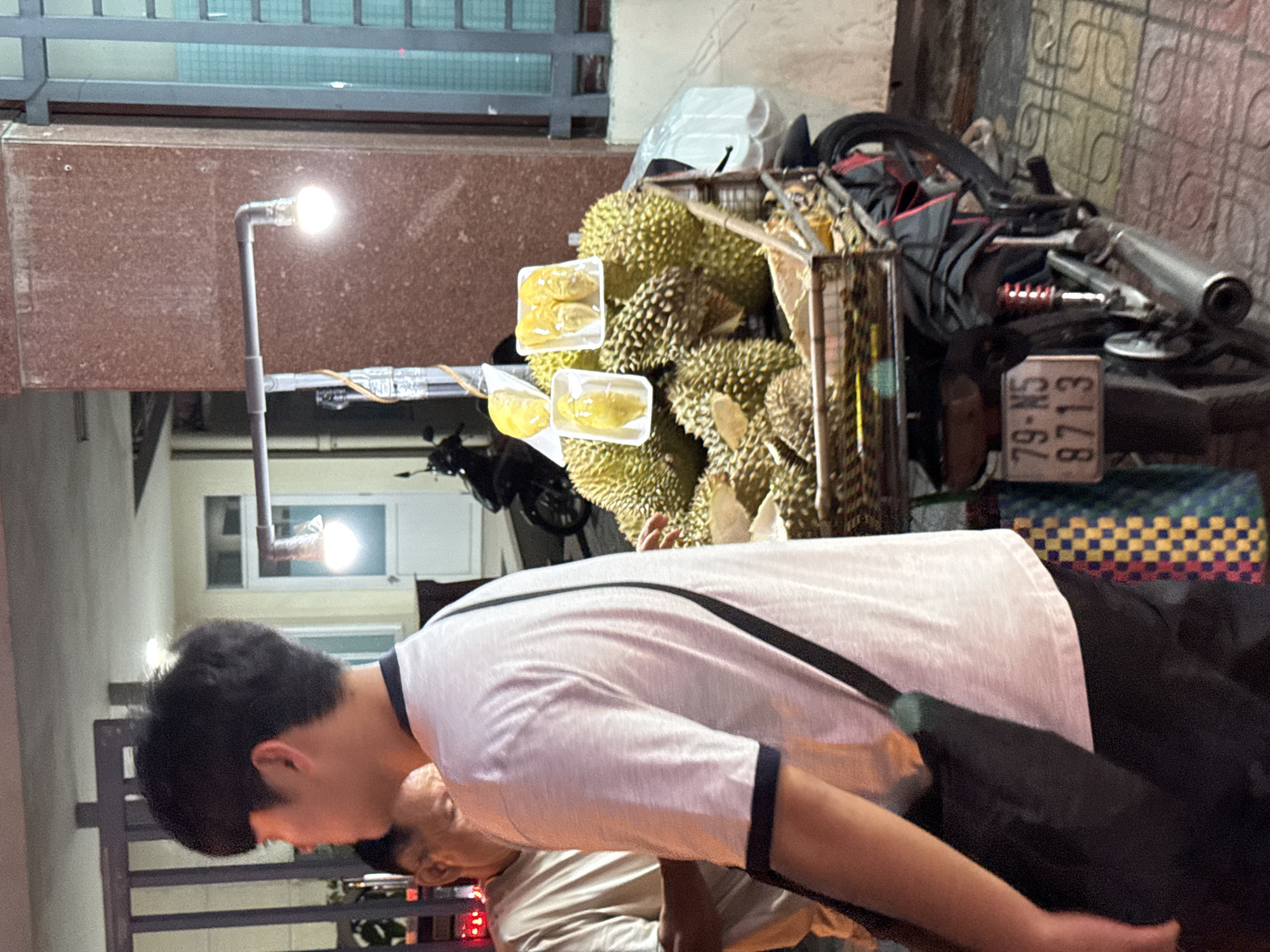 In Nha Trang, Vietnam, a young man with short dark hair and a white t-shirt with dark trim walks past a street vendor's stall loaded with durian fruit. The stall is mounted on the back of a parked motorcycle, which has a license plate reading "79-N5 8713". The durian is displayed in a basket and in clear plastic containers, some of which are topped with yellow fruit flesh. A second man, older with short dark hair, is partially visible behind the younger man, facing the durian stall. The scene is illuminated by two bright, exposed light bulbs suspended from a metal pole. The background features a building with a reddish-brown textured facade and a large window with blue bars. The ground is paved with tiles.