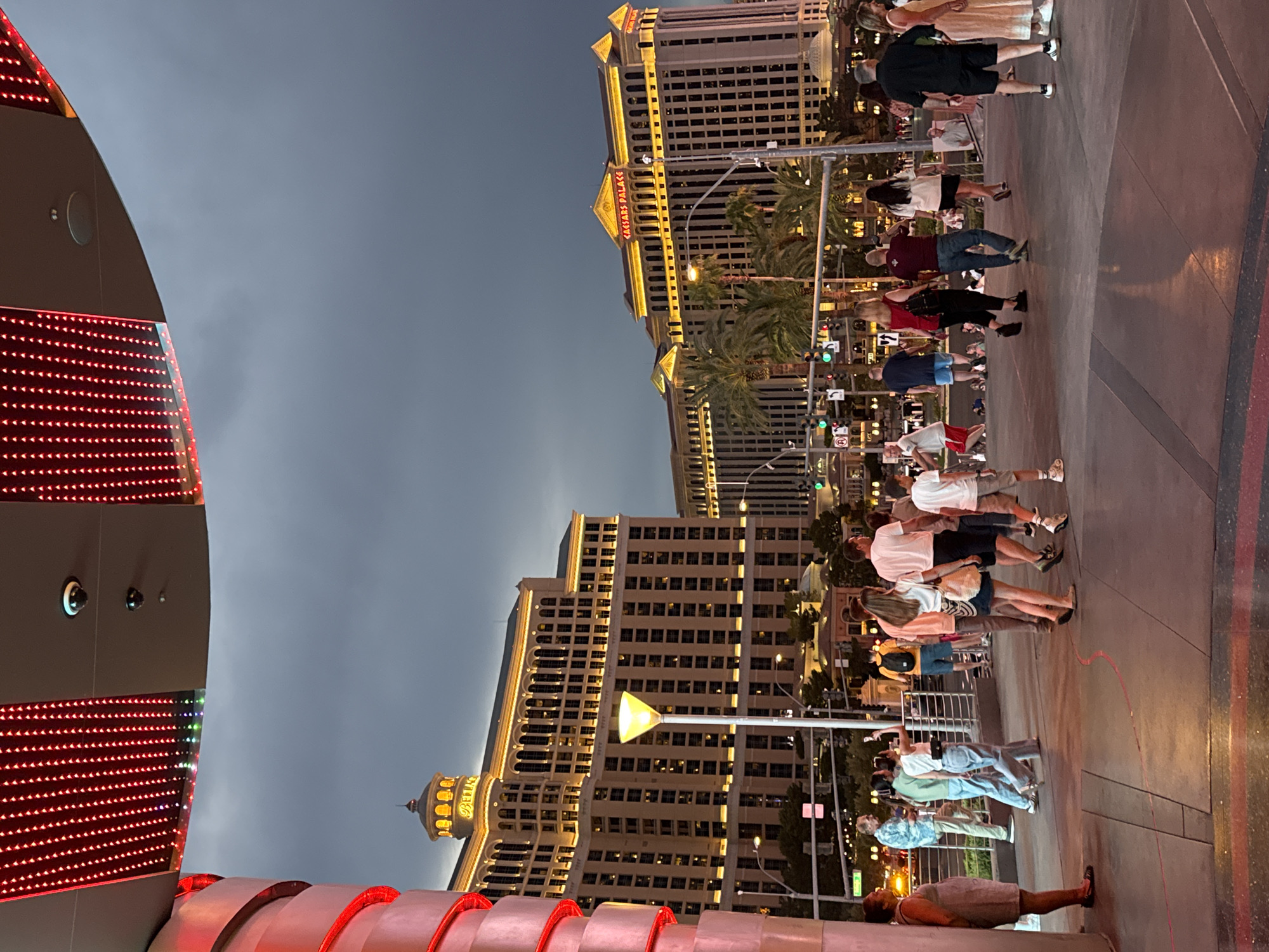 The image captures a twilight scene on the Las Vegas Strip in Paradise, Nevada, with the iconic Bellagio and Caesars Palace hotels illuminated in the background. A group of pedestrians is walking on a paved walkway under a dark, cloudy sky. In the foreground, a red, cylindrical structure with spiral detailing and overhead LED lights is partially visible on the left. A street lamp with a yellow light fixture stands near the Bellagio hotel. The walkway has a reddish-pink line marking a boundary. People are dressed in casual summer attire, with some individuals holding phones and bags. The lighting is a mix of the ambient twilight and the artificial lights from the buildings and street lamps.