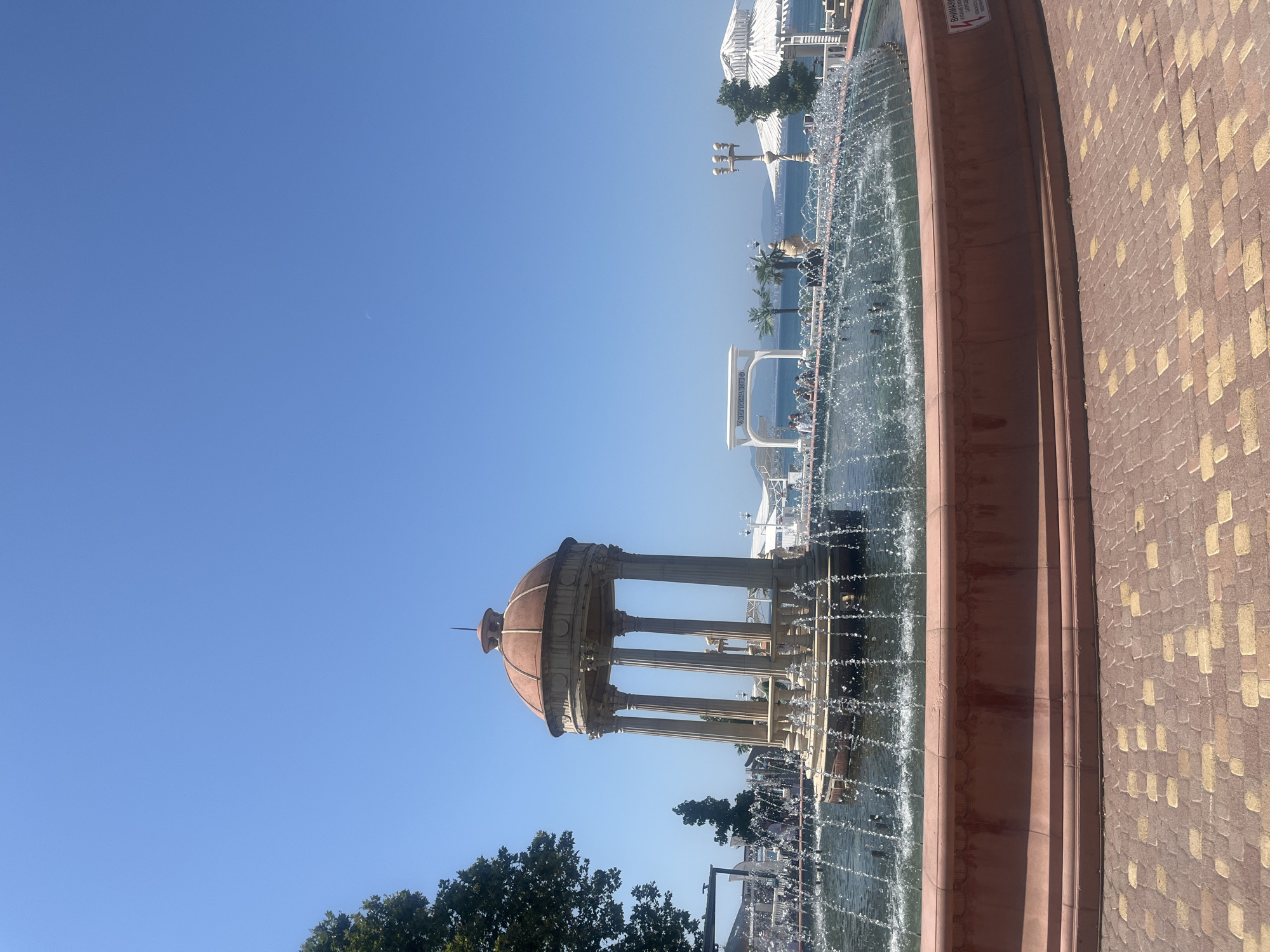 This image captures a scene in Novorossiysk, Russia, featuring a large, ornate fountain in the foreground with water spraying in arcs. The fountain is made of stone and has a domed roof supported by columns. Beyond the fountain, a clear blue sky stretches overhead. In the middle ground, there is a paved area with people walking and sitting. A white archway with text visible, palm trees, and white structures that appear to be gazebos or shelters are also present. The background shows a body of water, likely the Black Sea, with a coastline and buildings visible in the distance. The ground in the immediate foreground is paved with interlocking bricks in shades of red and yellow. A red sign with Cyrillic text is affixed to the side of the fountain's basin.