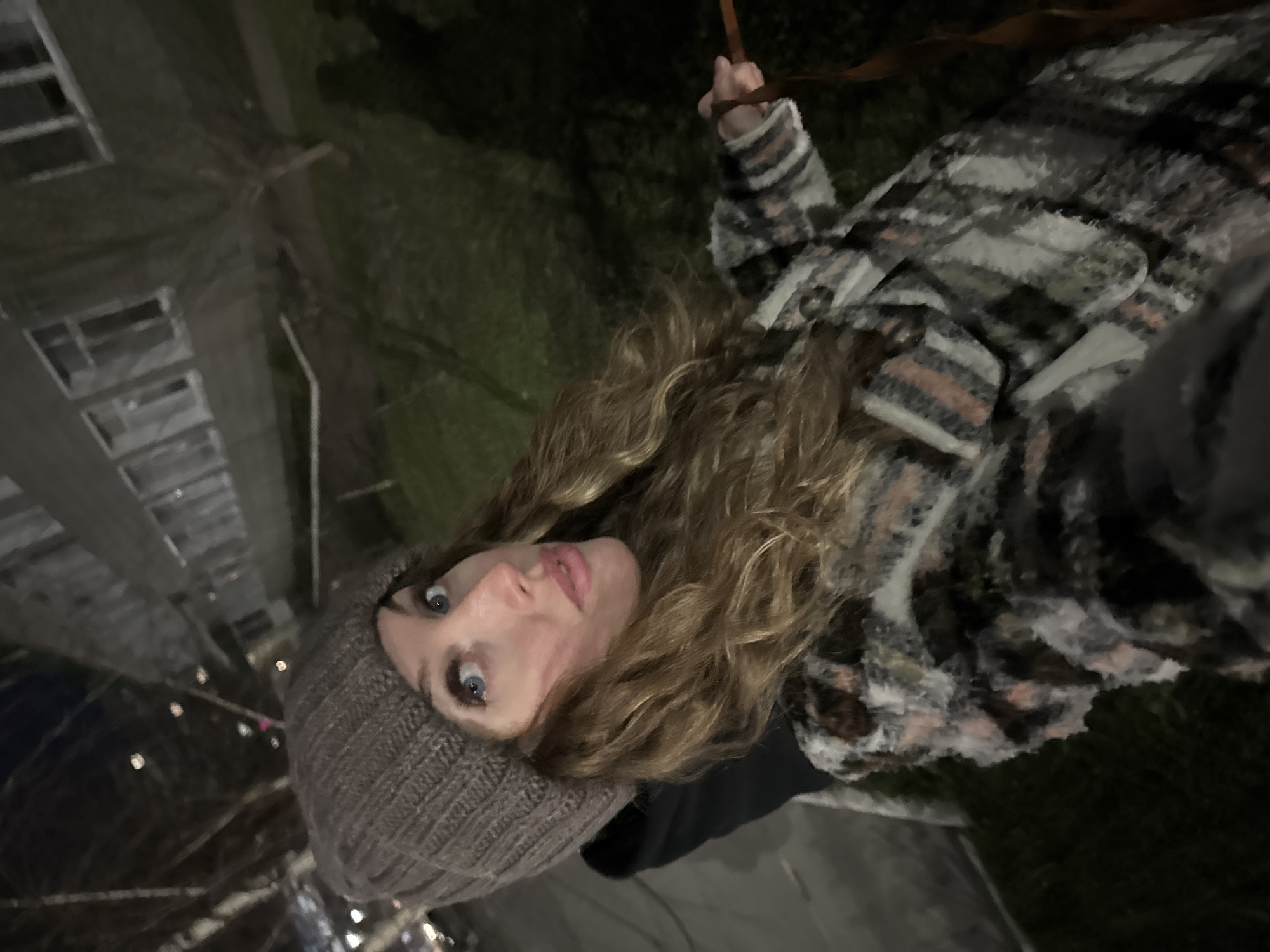 A woman with long, light brown curly hair and blue eyes is posing for a selfie. She is wearing a brown knit beanie and a plaid jacket. The background shows a tall building with multiple floors and windows, illuminated by artificial lights. It appears to be evening or nighttime. The location may be Chaikovsky, Russia.