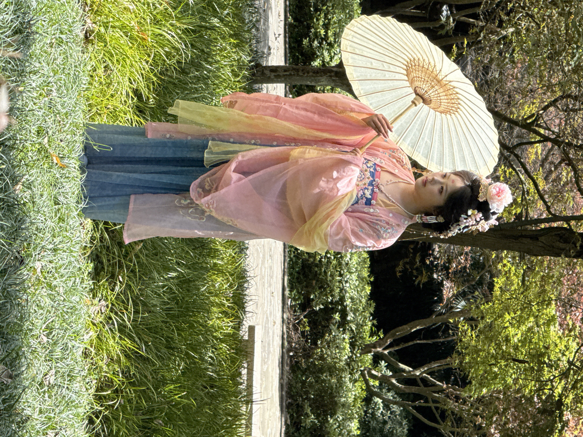 A portrait shows a person in Yuzhong District, China, lying on the ground next to a concrete path, with tall grass growing to the left of the path and a green leafy bush to the right. The individual wears a long, flowing dress with a blue base and a pink and yellow overlay, adorned with floral accents and necklaces. The person holds a cream-colored parasol above, while an ornate floral headpiece adorns their dark hair. Trees with green and brown leaves are in the background.