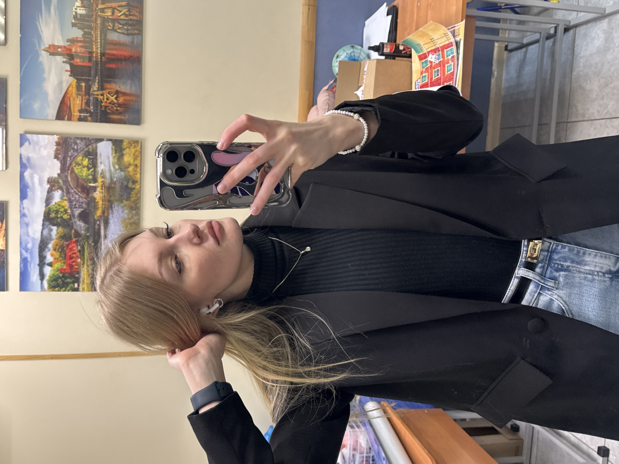 The image depicts a young woman taking a mirror selfie indoors. She has long, straight blonde hair and is shown from the chest up, looking towards the camera reflected in the mirror. She wears a black ribbed turtleneck sweater, a black blazer, and light blue jeans with a visible gold buckle or zipper detail on the waistband. Her accessories include a silver necklace with a small clear gem, a white beaded bracelet with two silver embellishments on her right wrist, and a black smartwatch on her left wrist. White wireless earbuds are in her ears. Her smartphone, held in her right hand and covered by a clear case with abstract grey and purple shapes, is positioned to capture her reflection. Her left hand is gently touching her hair near her ear.
The background features a light-colored wall adorned with several framed art prints or photographs, depicting landscapes and cityscapes. Notable among these are a city skyline reflected in water and a stone bridge over a river with autumn-colored trees. To the right, a wooden desk or shelf is cluttered with various items, including a blue globe, a brown cardboard box, papers, and a colorful poster with architectural drawings. A metal shelving unit is partially visible further back.
The woman's expression is neutral, with her lips slightly parted. The setting appears to be an indoor space, possibly an office, classroom, or personal studio, given the mix of decorative art and functional workspace items. There is no legible text visible on any objects in the scene. The time of day and weather are not discernible from the indoor lighting. The overall scene suggests a casual environment in Perm, Russia, as per the provided context.
