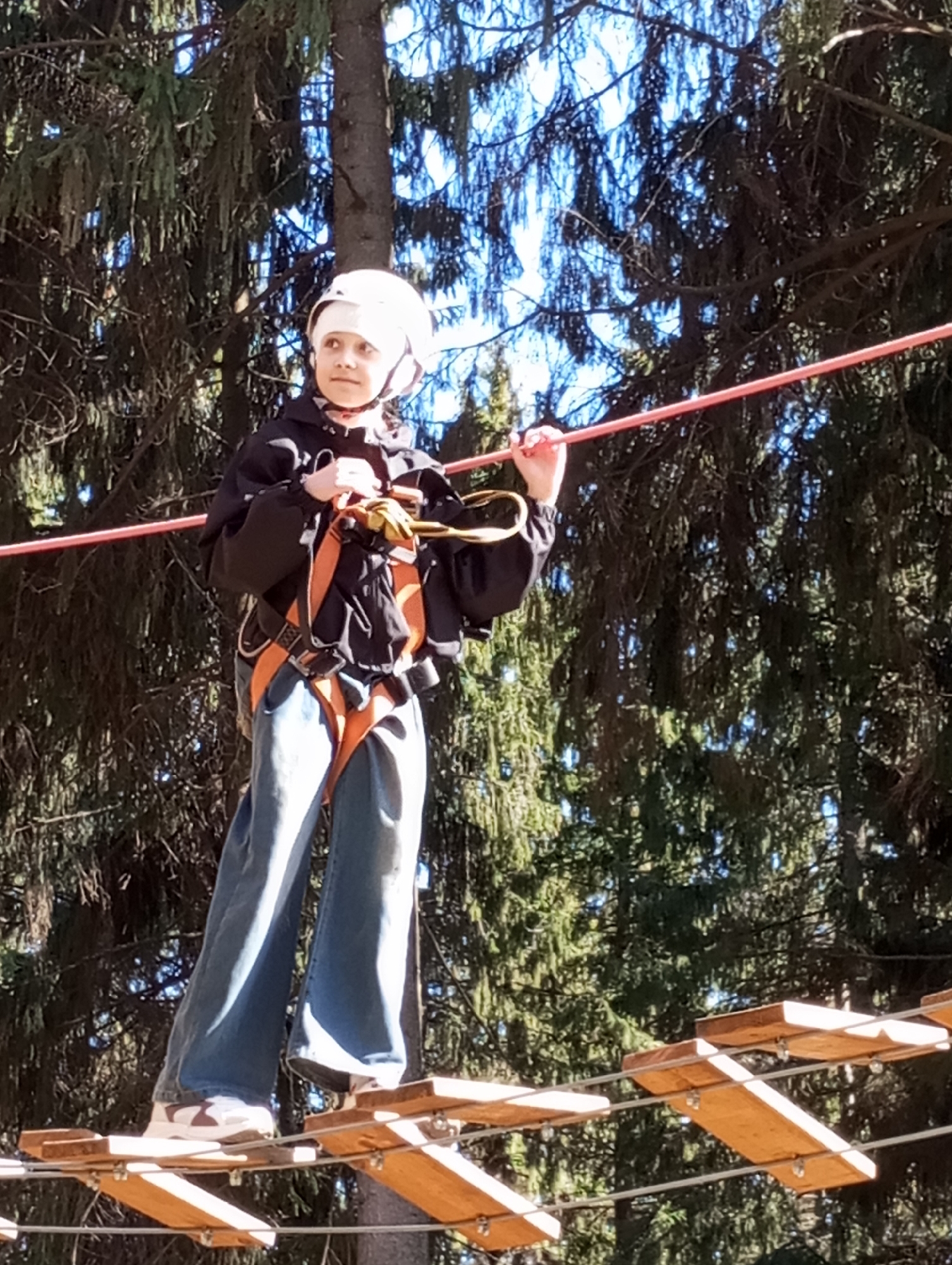 A child wearing a white helmet and safety harness is carefully walking across a wooden rope bridge in what appears to be a high-ropes course. The child is wearing a dark jacket and blue jeans, with white sneakers visible. The background shows a dense forest setting with tall evergreen trees. Based on context clues and the description, it is likely that this is an outdoor recreation area in Izhevsk, Russia.