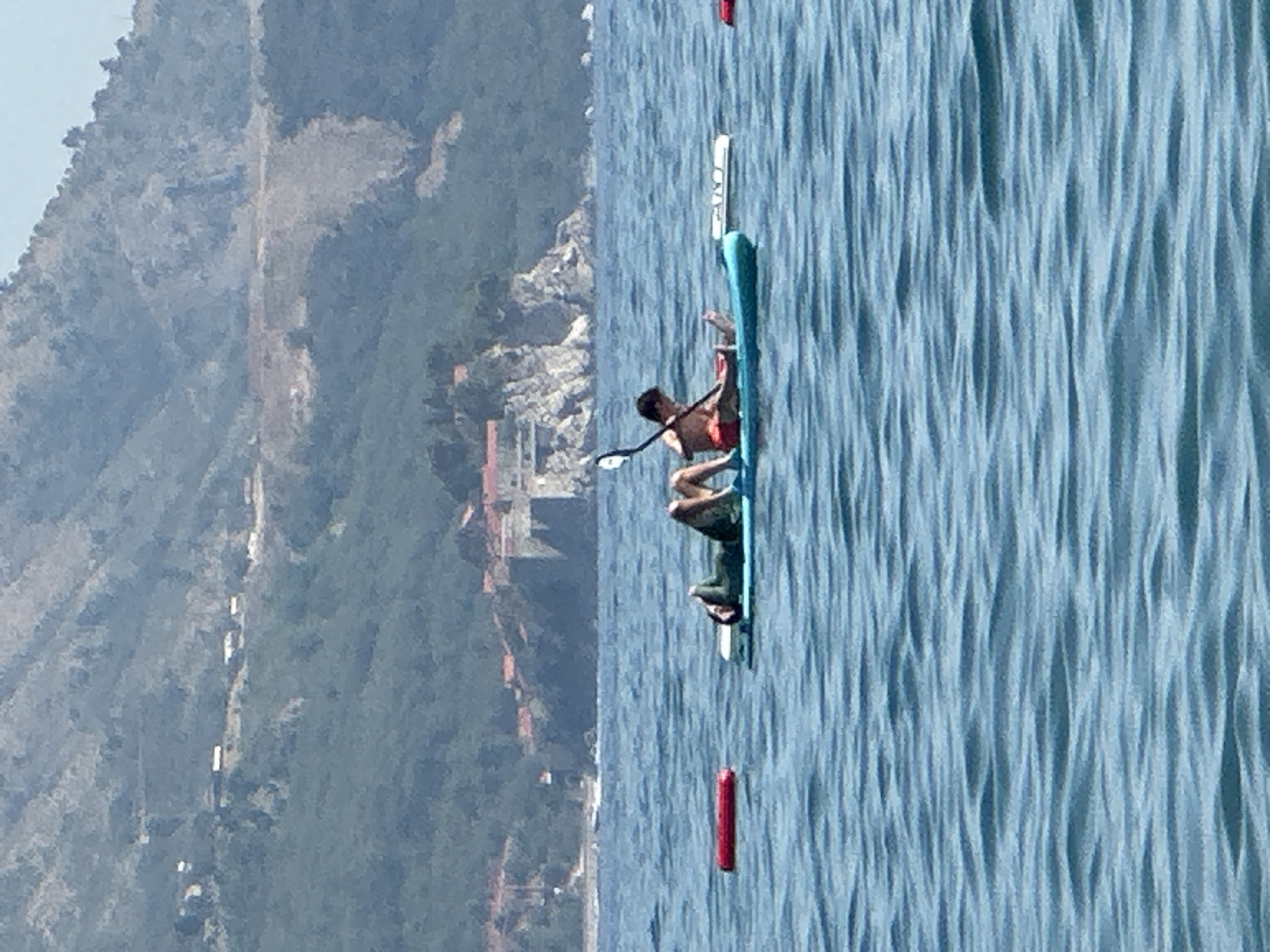 In the waters off the coast of Boreti, Montenegro, two individuals are engaged in recreational activity on a turquoise stand-up paddleboard. One person, wearing a dark green top and red shorts, is lying down on their back. The other person, wearing red swim trunks, is seated and holding a paddle. A red inflatable buoy is visible in the water to the left of the paddleboard, and another white object, possibly a cooler or another float, is to the right. In the background, the hilly coastline of Boreti is dotted with buildings, and the sea displays gentle ripples.