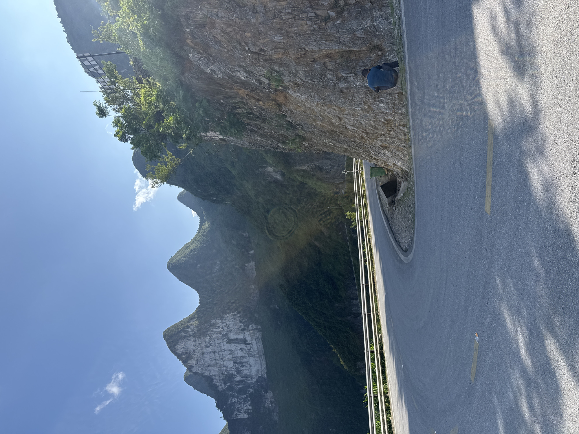 A road cuts through a mountainous landscape in Đồng Văn Commune, Vietnam. The road, marked with a yellow line, curves along the edge of a steep rock face. A metal barrier runs along the edge of the road, separating it from the drop-off. Further up the mountain are dense forests and rocky peaks under a clear blue sky. A dark figure is visible perched on the side of the rock face, close to the road.