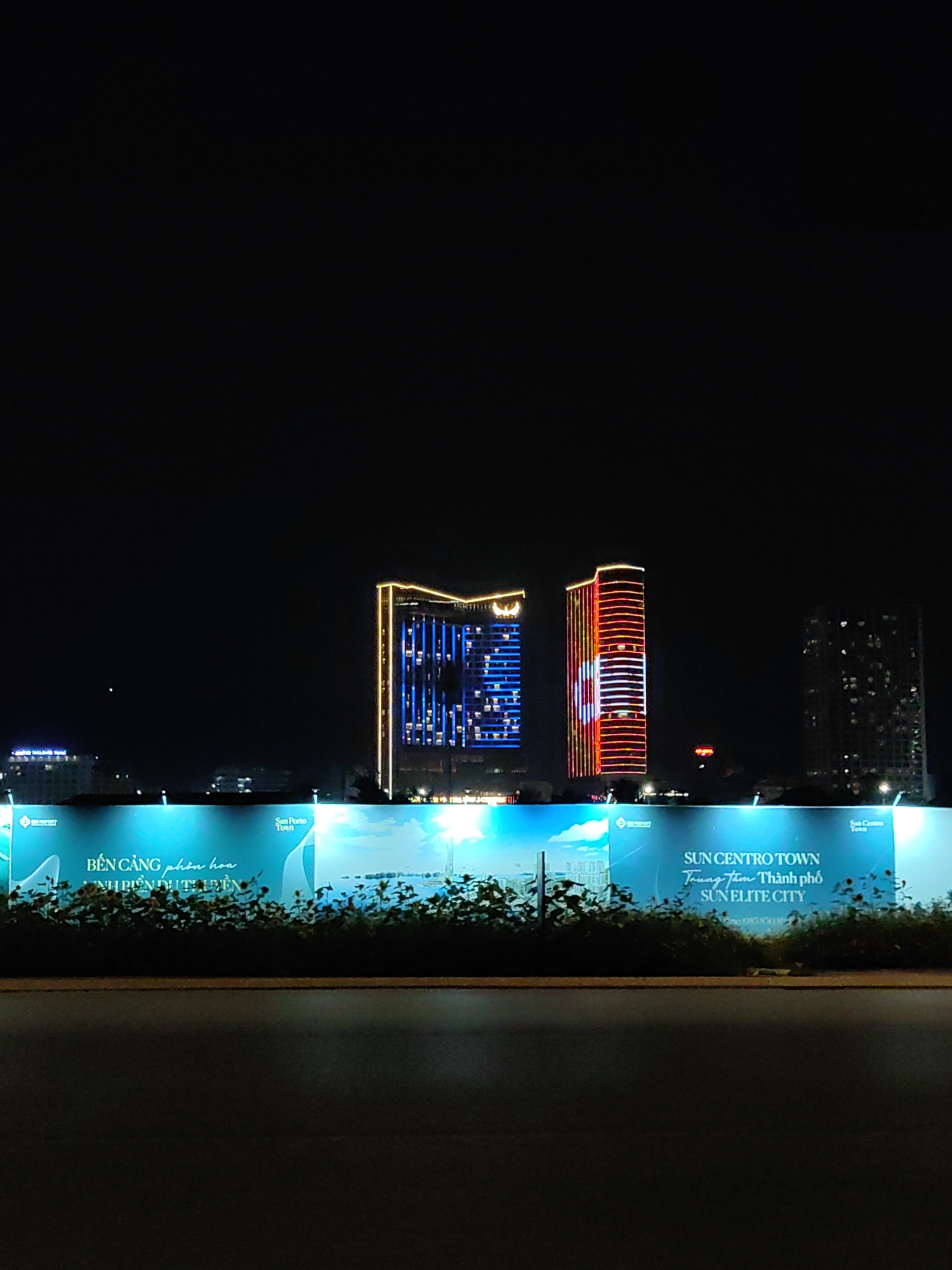 At night, the skyline of Bai Chay Ward, Vietnam, is visible beyond a low wall or barrier and some greenery. Three buildings stand out: one with blue lights, another with red lights, and a third, taller one to the right, with visible window lights. A large advertisement sign, brightly lit, sits in front of the greenery and the barrier. The sign contains the words "BEN CANG phon hoa", "ANH BIEN DUT UYEN", "SUN CENTRO TOWN", "Trung tam Thanh pho", and "SUN ELITE CITY". A road is visible in the foreground.