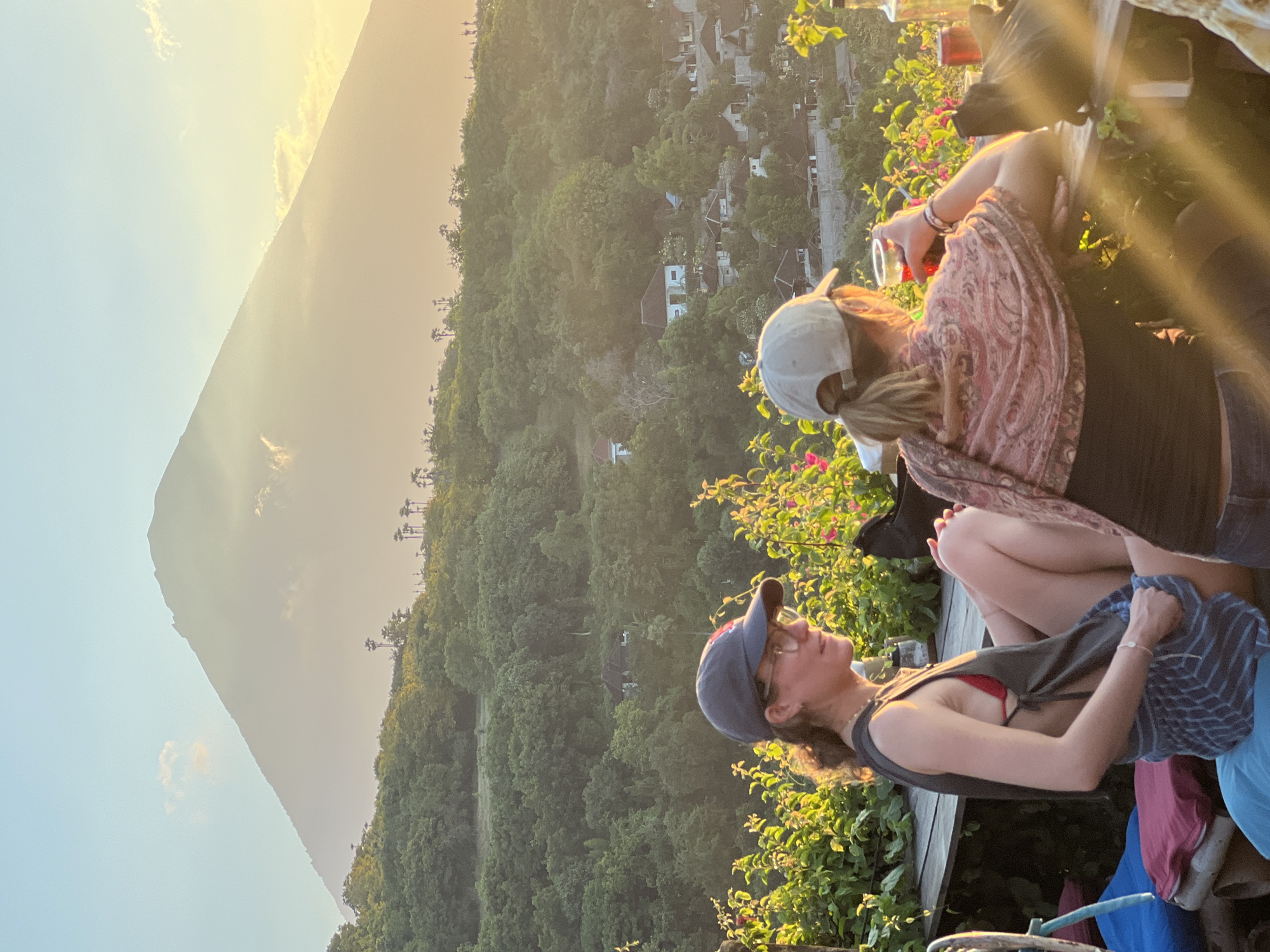 Two women are seated at a table outdoors, with a lush green hillside and a large, mist-shrouded mountain in the background. The woman on the left is wearing a dark sleeveless shirt over a red bikini top, blue and white striped shorts, a dark baseball cap, and glasses. She is looking to her right. The woman on the right is wearing a black tank top, a patterned pink scarf, and a light-colored baseball cap. She is holding a red drink and looking towards the mountain. The table has drinks and a bottle on it. The scene appears to be in Abang, Indonesia, given the presence of a conical volcano in the distance, characteristic of the region. The lighting suggests it is either sunrise or sunset.