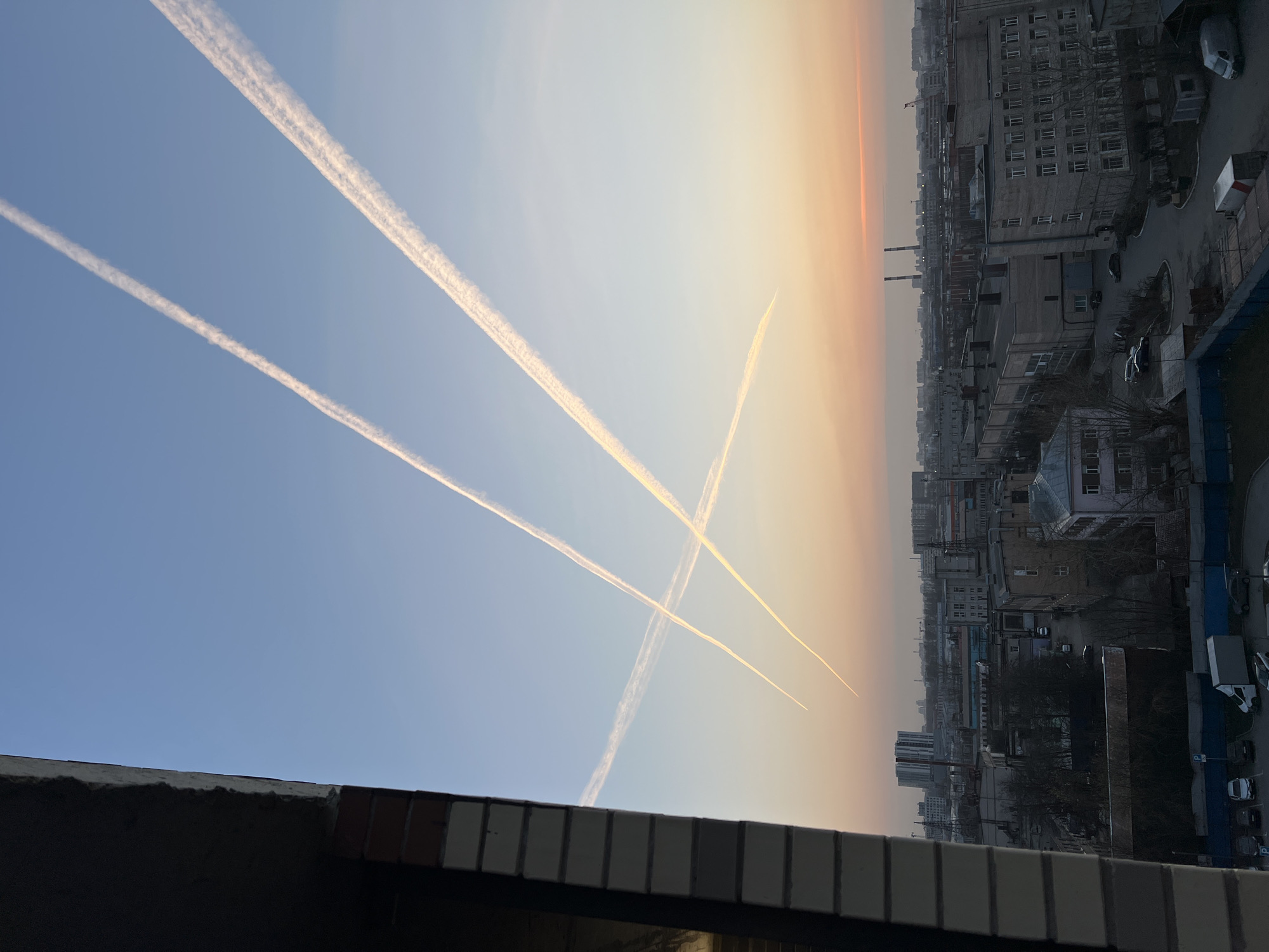 The photograph, likely taken from a high vantage point in Saint Petersburg, Russia, captures an urban scene under a sky painted with the hues of dawn or dusk. Buildings of various sizes and architectural styles form the cityscape, appearing dense and grey. The sky is layered with color, transitioning from a light blue to a warm orange near the horizon. Several contrails from airplanes crisscross the sky, their white lines contrasting against the sky's gradient. In the foreground, a portion of a building's rooftop or balcony is visible, constructed with a combination of brick and concrete.