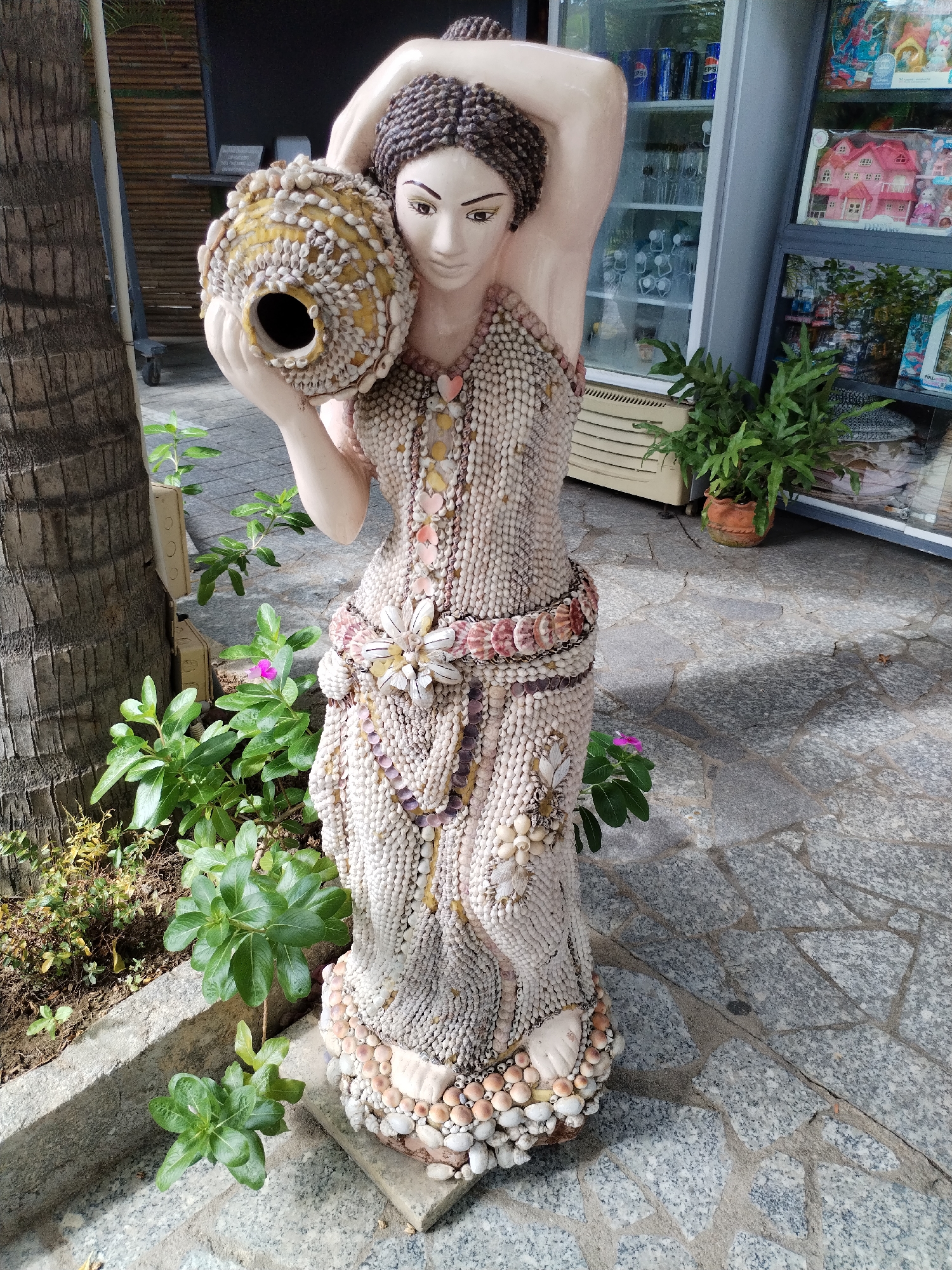 The image shows a decorative statue of a woman made of shells. She is holding a shell-covered pot on her shoulder. The statue is standing on a paved surface near a tree and some plants. In the background, a building with a glass display case filled with various items is visible. The statue is located in Bac Nha Trang Ward, Vietnam.