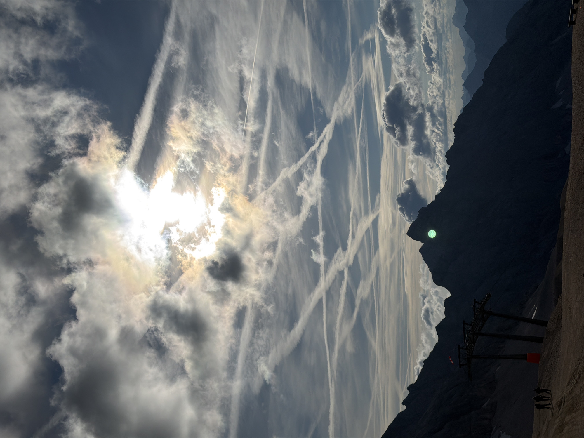 This image depicts a panoramic view of a mountainous landscape, likely the Alps, near Garmisch-Partenkirchen, Germany. The foreground features a dirt path and the silhouettes of several large, dark mountain peaks. In the mid-ground, a set of imposing, dark ski lift towers stands prominently. Three figures, also in silhouette, are walking on the path near the ski lift. The sky is dramatic, filled with a mix of dark, heavy clouds and lighter, wispy clouds. Sunlight breaks through the clouds, creating bright highlights and a hazy glow, with numerous contrails crisscrossing the sky. The overall impression is one of a vast, rugged, and somewhat atmospheric natural environment.